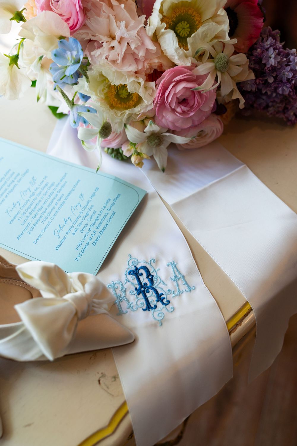 A romantic wedding detail shot with fresh pink and white flowers next to a blue invitation card and monogrammed napkin.