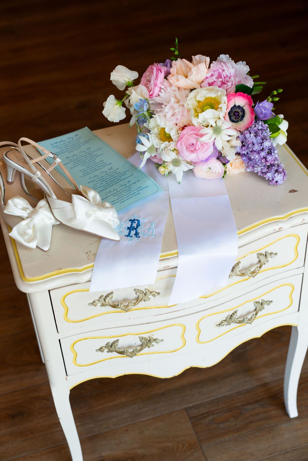Wedding accessories including bridal shoes and bouquet arranged on an ornate white vintage dresser.