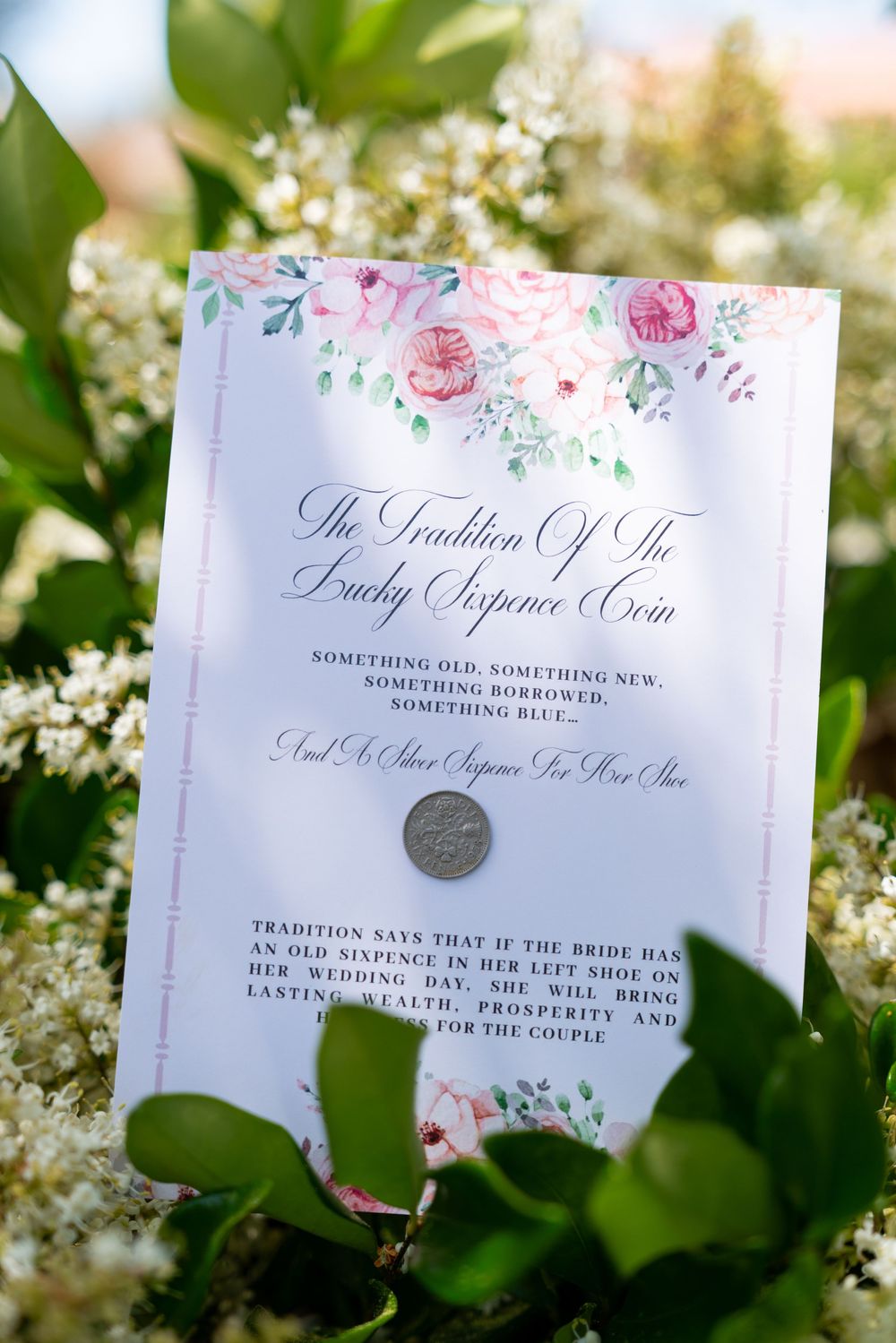 A wedding tradition card with delicate pink floral design and calligraphy text placed among white flowers outdoors.