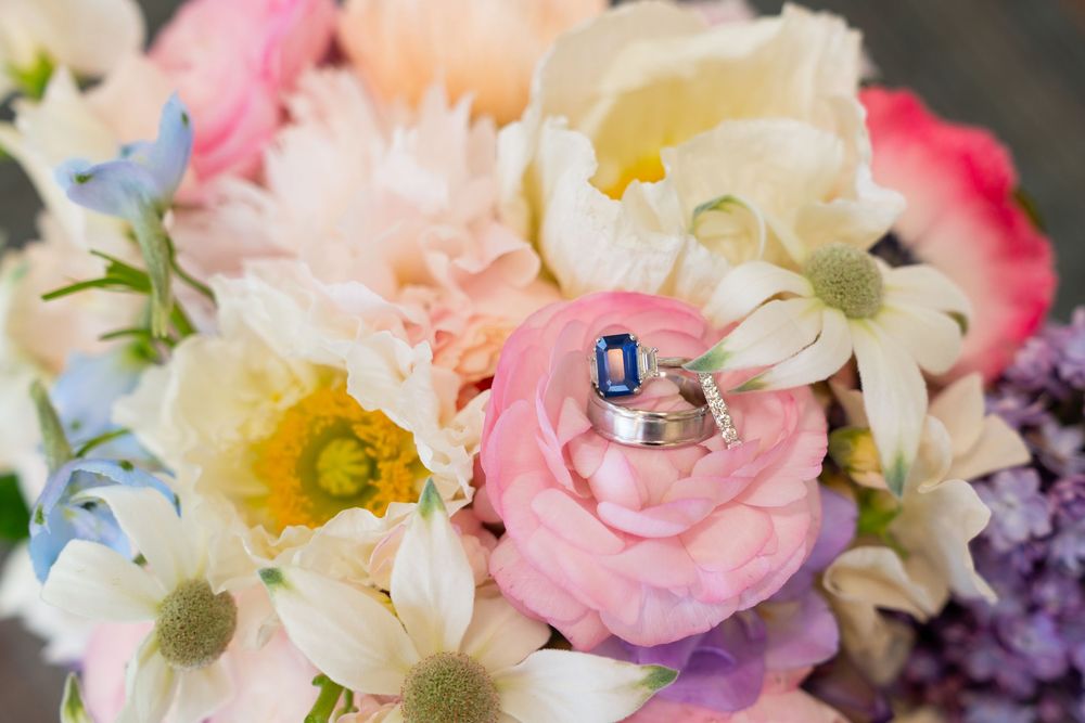 Two elegant wedding rings nestled in a pastel bouquet of pink roses, daisies and purple flowers in soft focus.