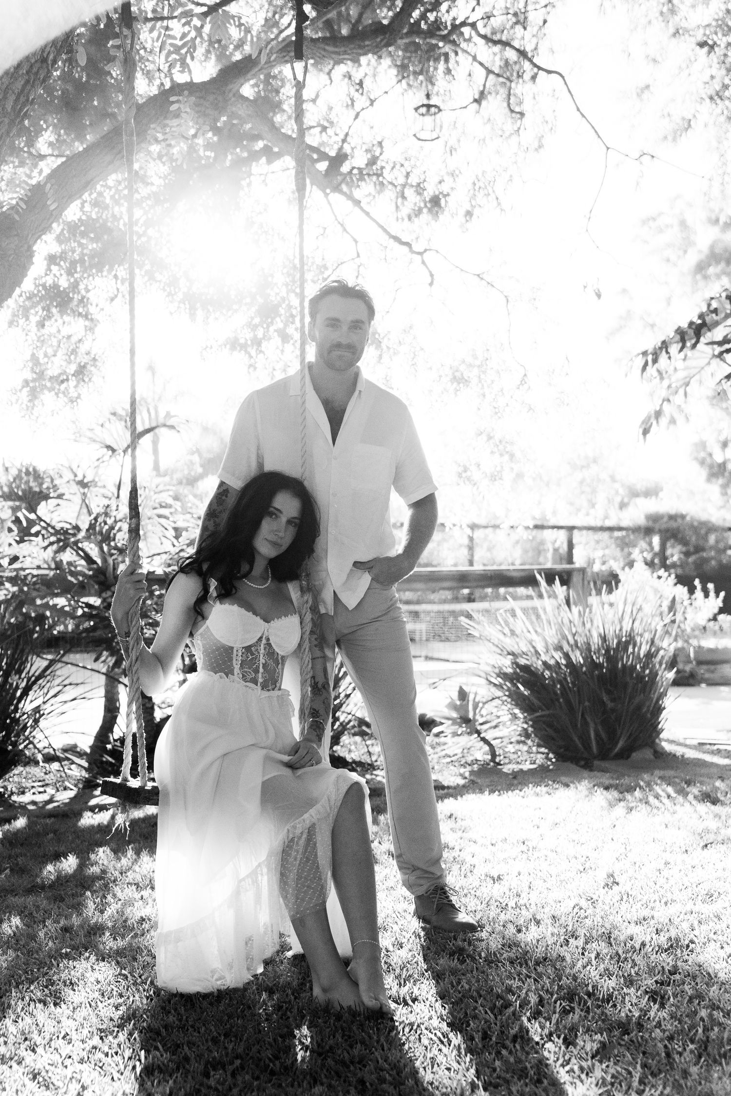 A black and white portrait taken outdoors under trees with dramatic natural lighting and shadows.