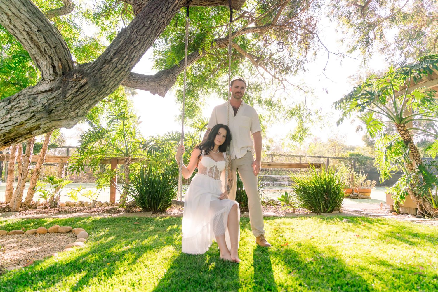 A couple stands together on a sunny lawn with trees and natural lighting creating a dreamy outdoor atmosphere.