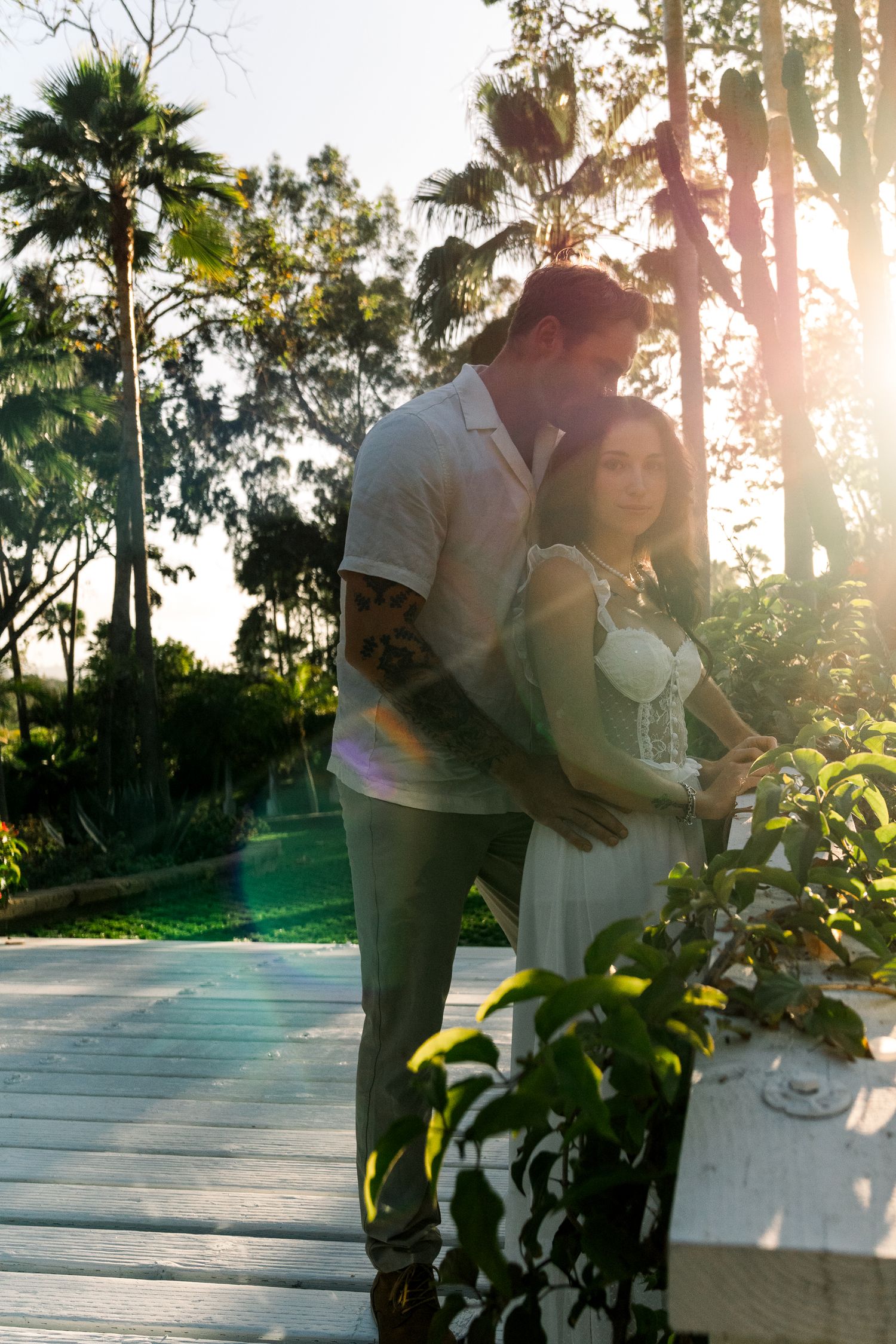 A couple embraces on a deck surrounded by palm trees and tropical foliage at sunset.