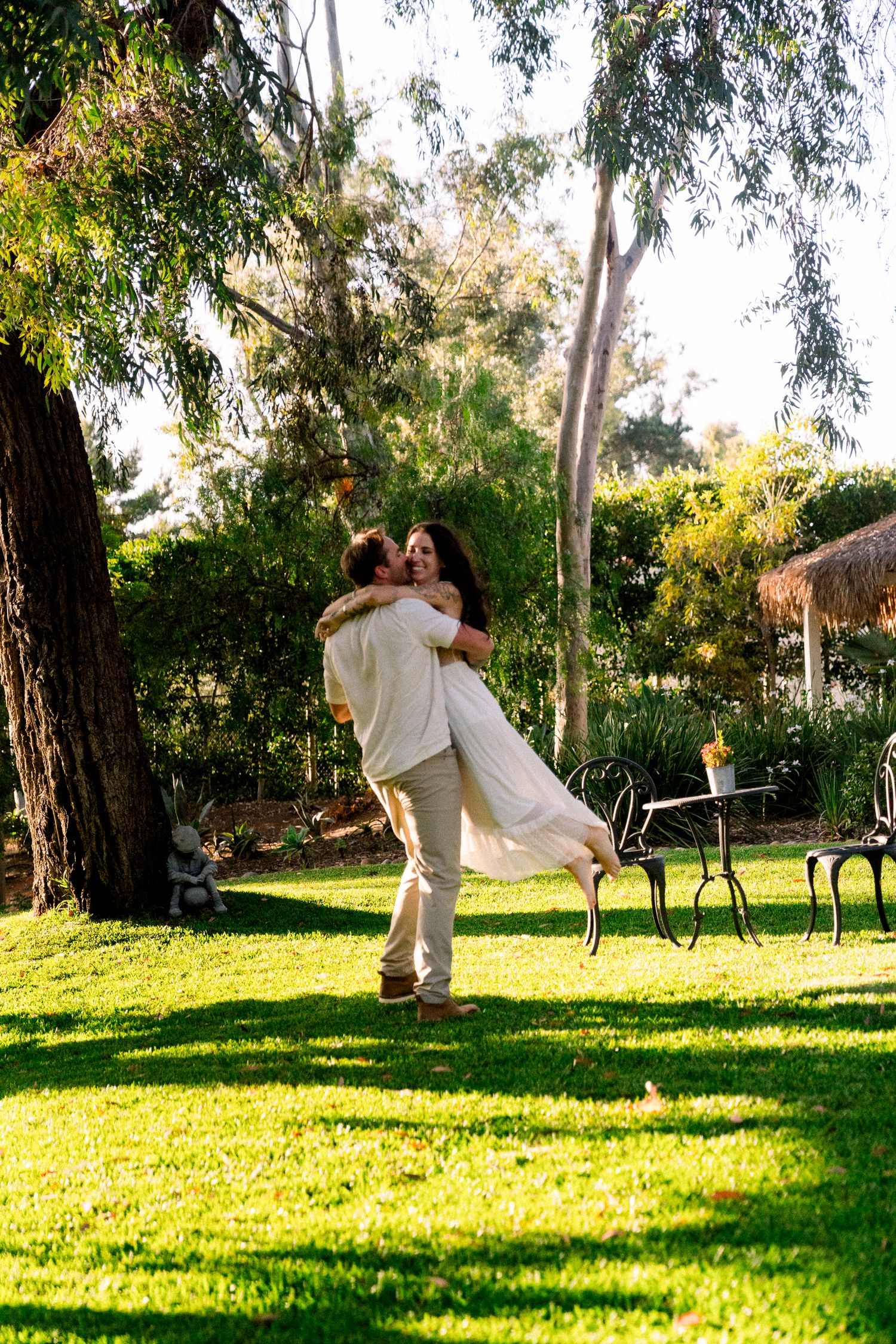 A joyous embrace between a couple in white attire on a sunlit garden lawn with trees in the background.