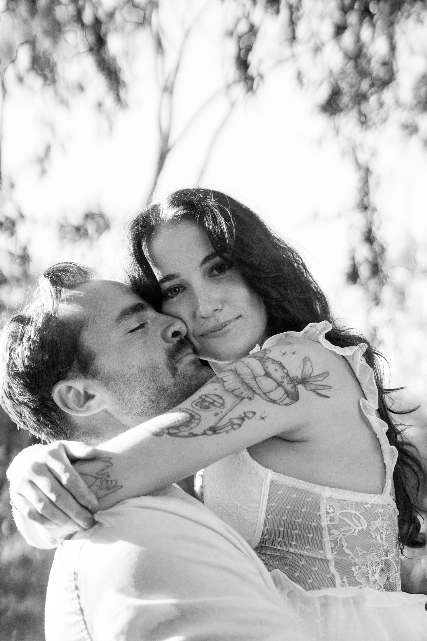 A romantic black and white portrait of a couple embracing outdoors with sunlit trees in the background.
