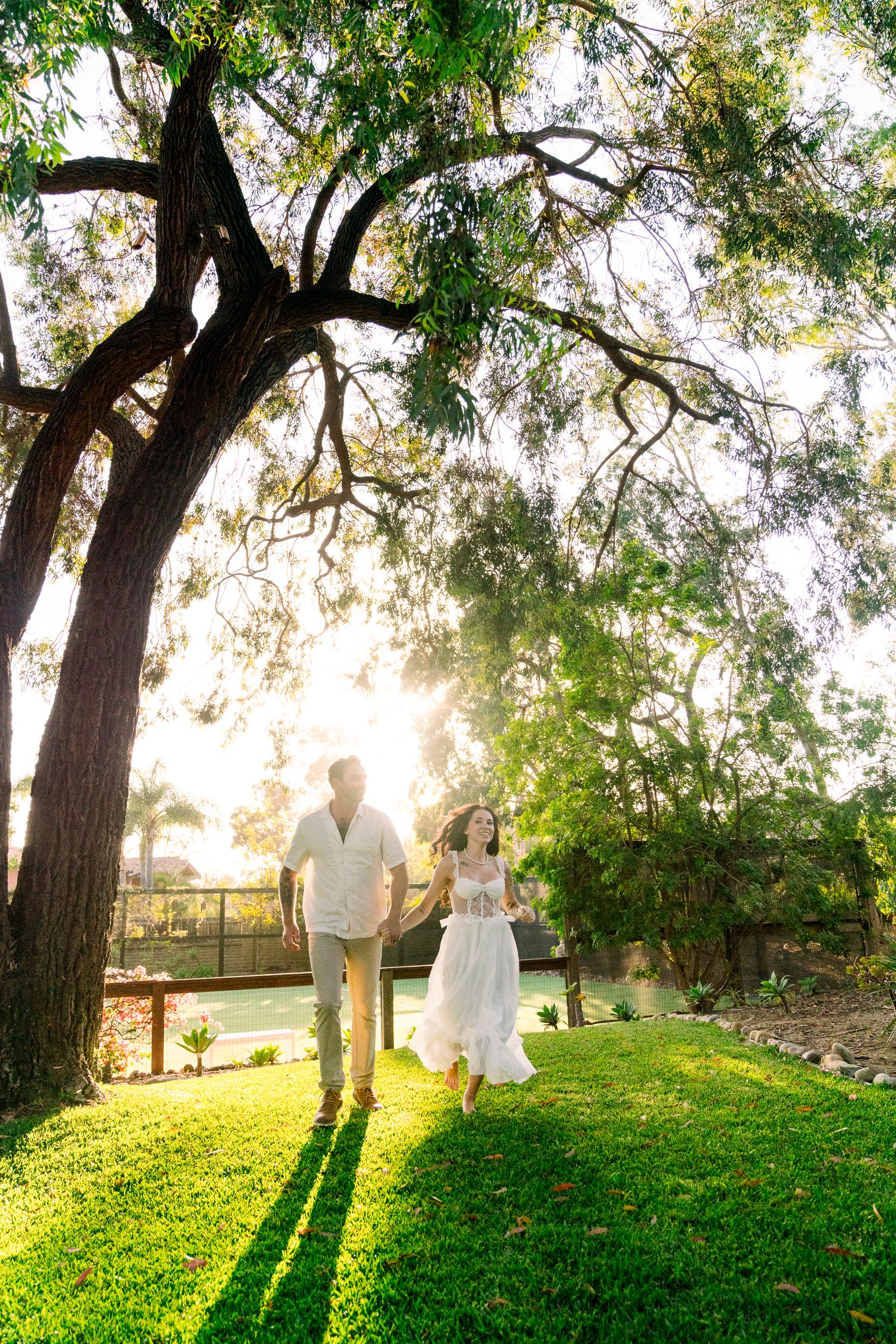 A romantic silhouette of a couple standing on grass under tall trees at sunset with golden light filtering through.