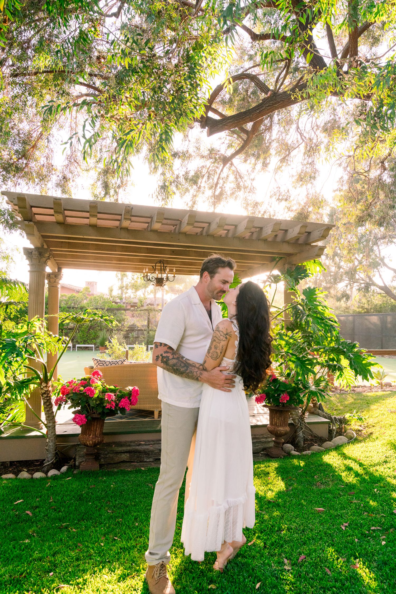 Romantic couple embraces under rustic pergola in lush garden setting with sunlit greenery and flowers.