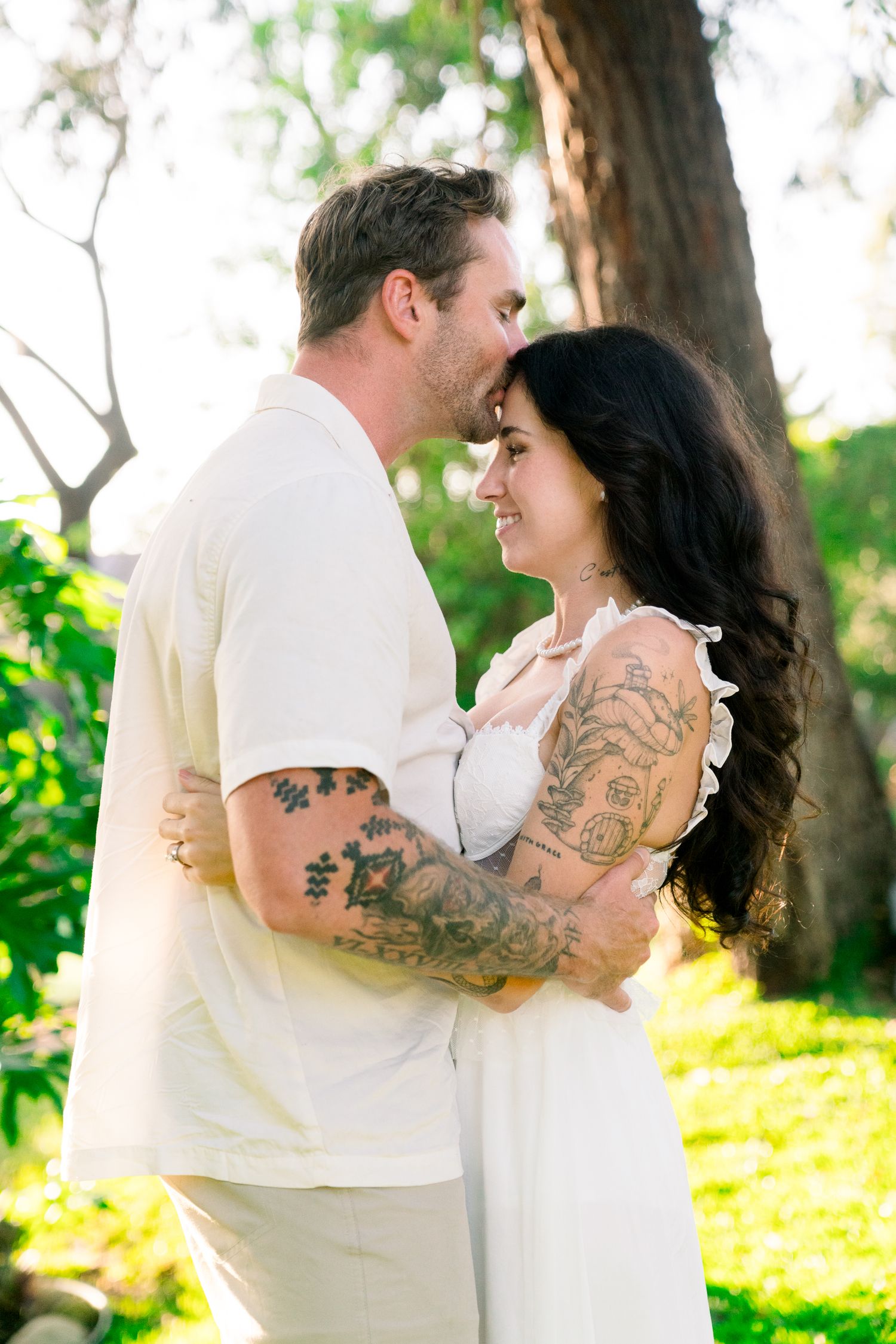 Romantic couple in white embracing outdoors in a garden setting with visible tattoos on their arms.