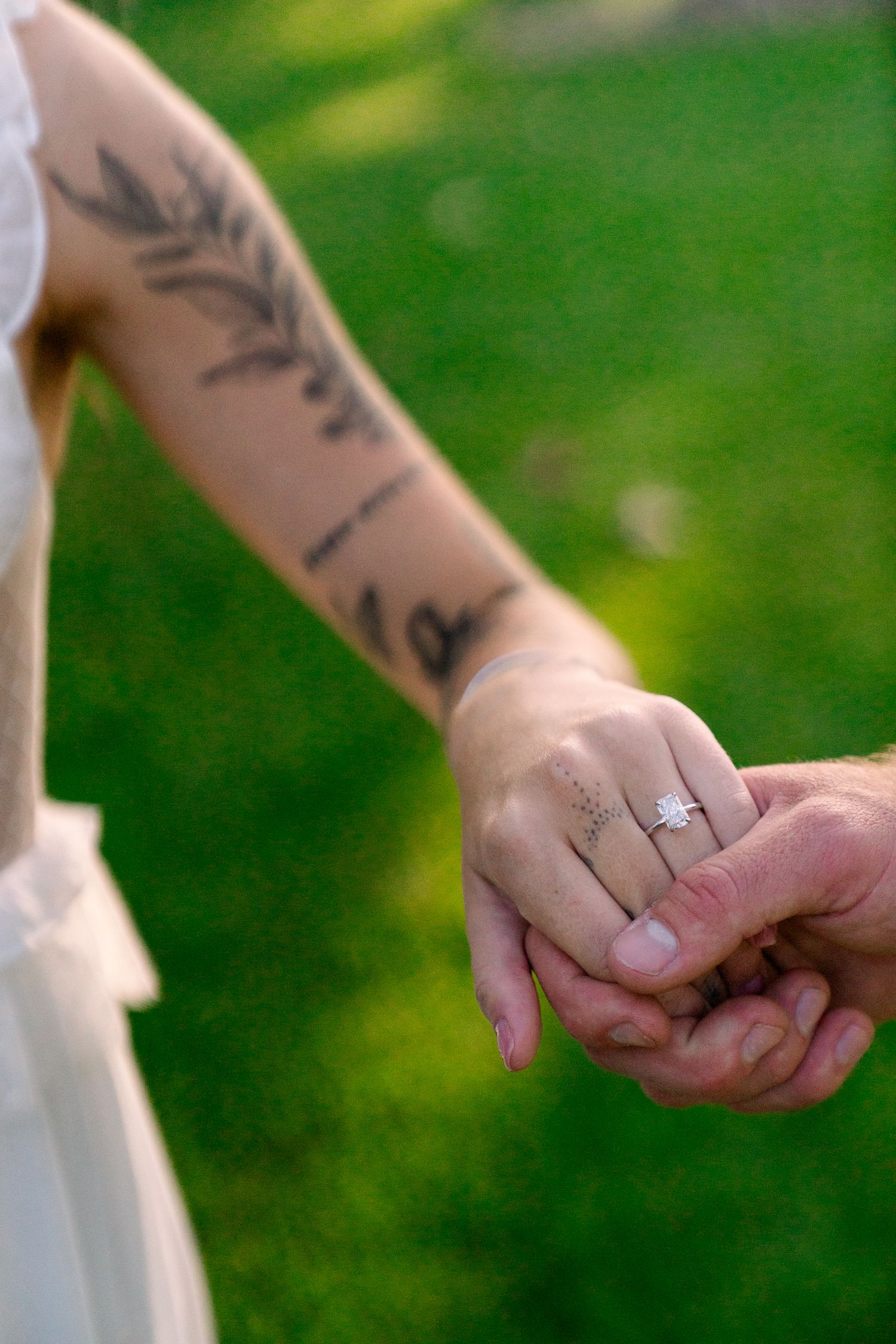 Close-up of intertwined hands showing a wedding ring and floral tattoo against a green lawn background.