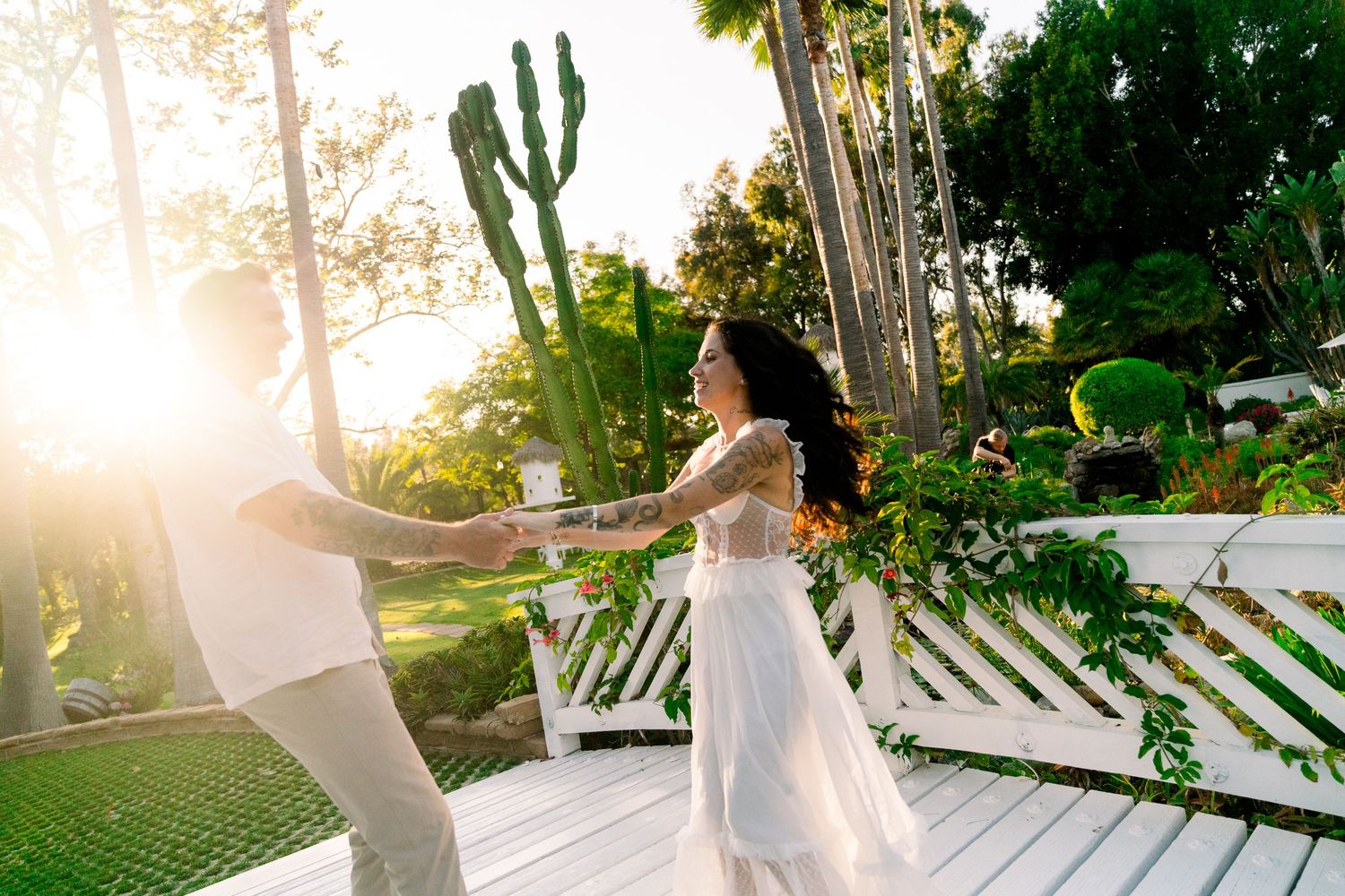 Two people dancing on a white deck with tropical plants and sunlight glowing in the background at sunset.