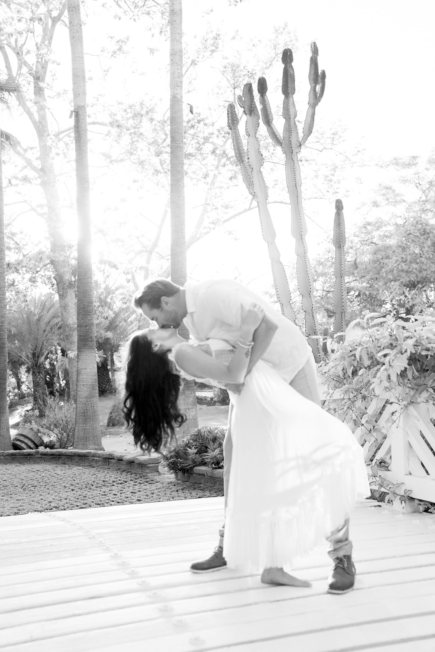 A black and white photo showing a romantic dance dip on an outdoor wooden deck with tall cacti in the background.
