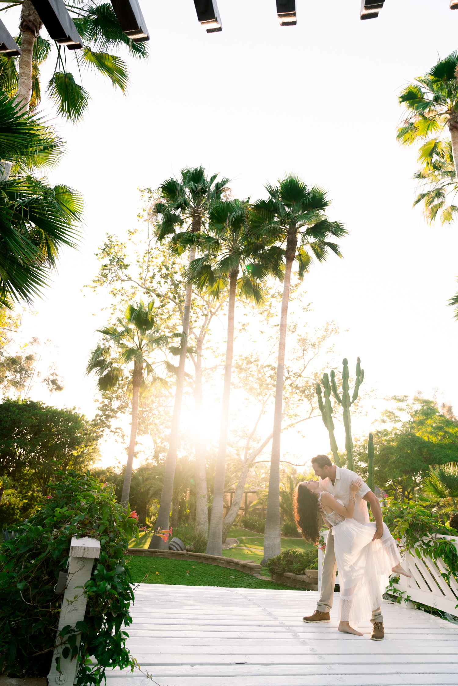 A romantic moment captured on a white bridge walkway with palm trees and golden sunlight filtering through.