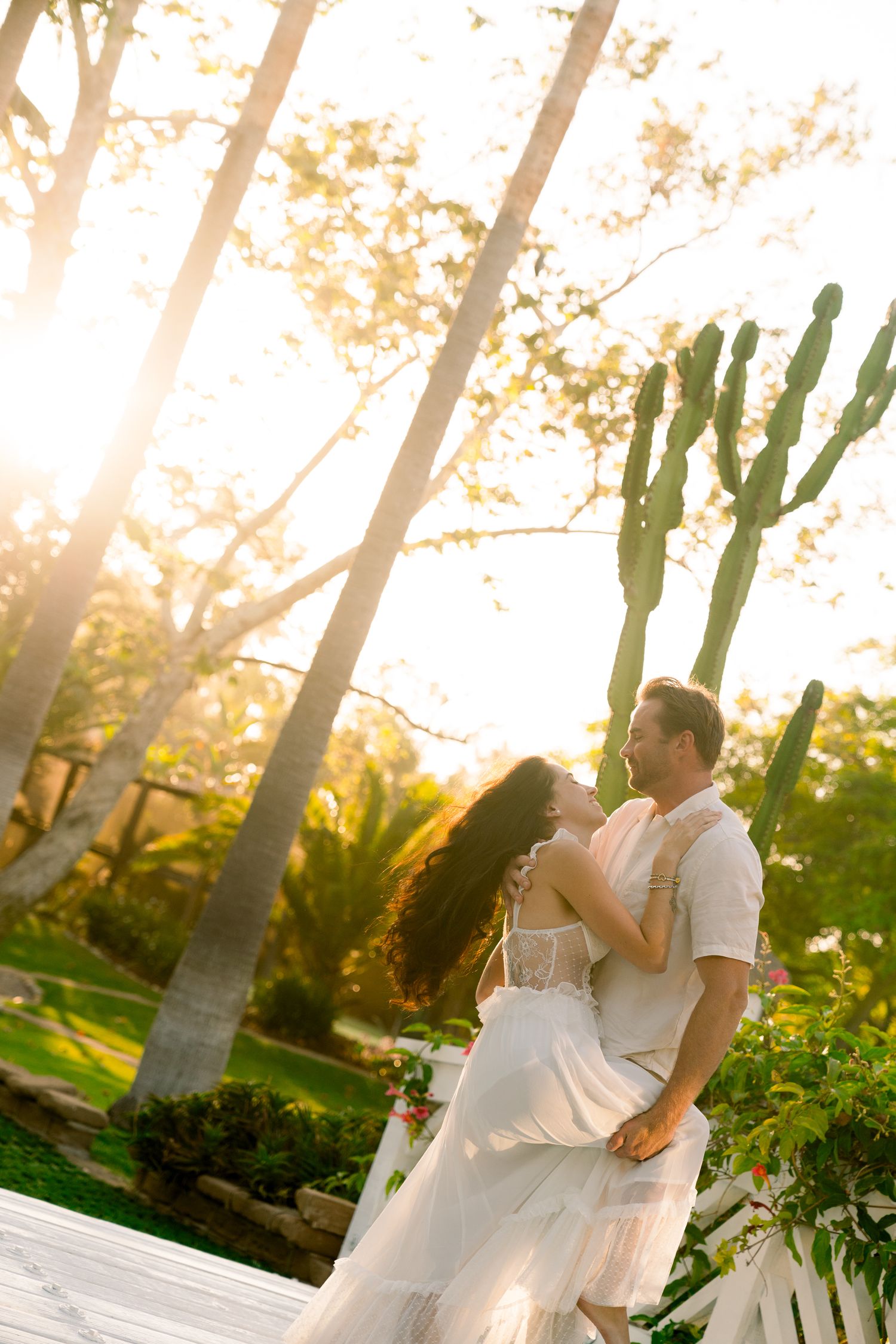 A romantic sunset wedding moment captured in a garden with tall palm trees and golden sunlight filtering through the foliage.