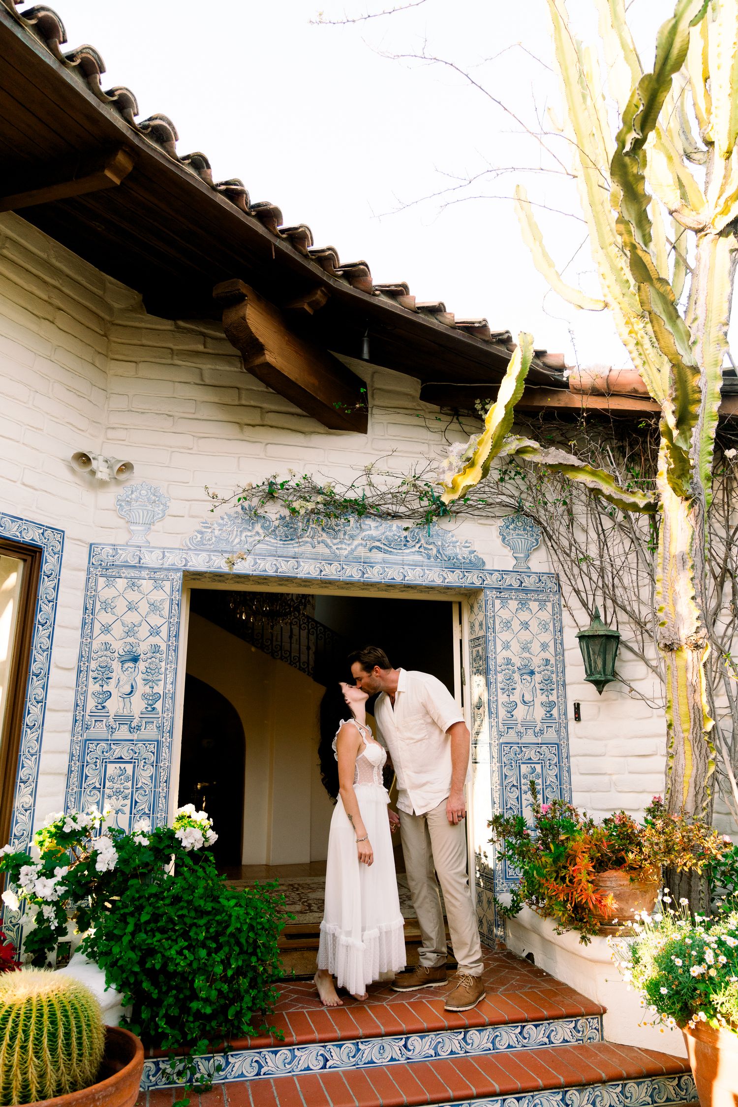 A couple embraces in a doorway of a Spanish-style building with blue tile work and potted desert plants.