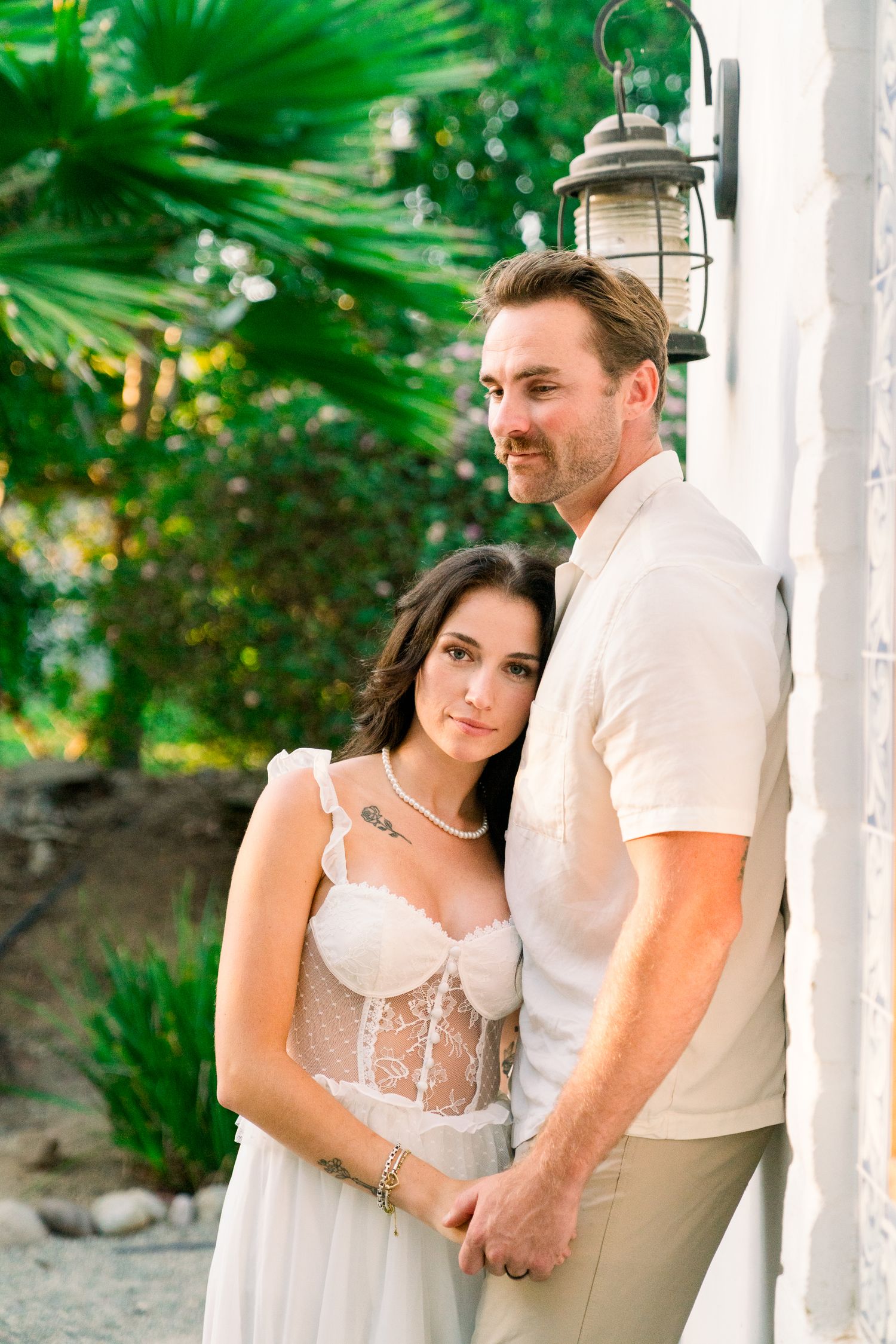 A romantic couple poses outdoors in white attire against a tropical garden backdrop with a decorative lantern.
