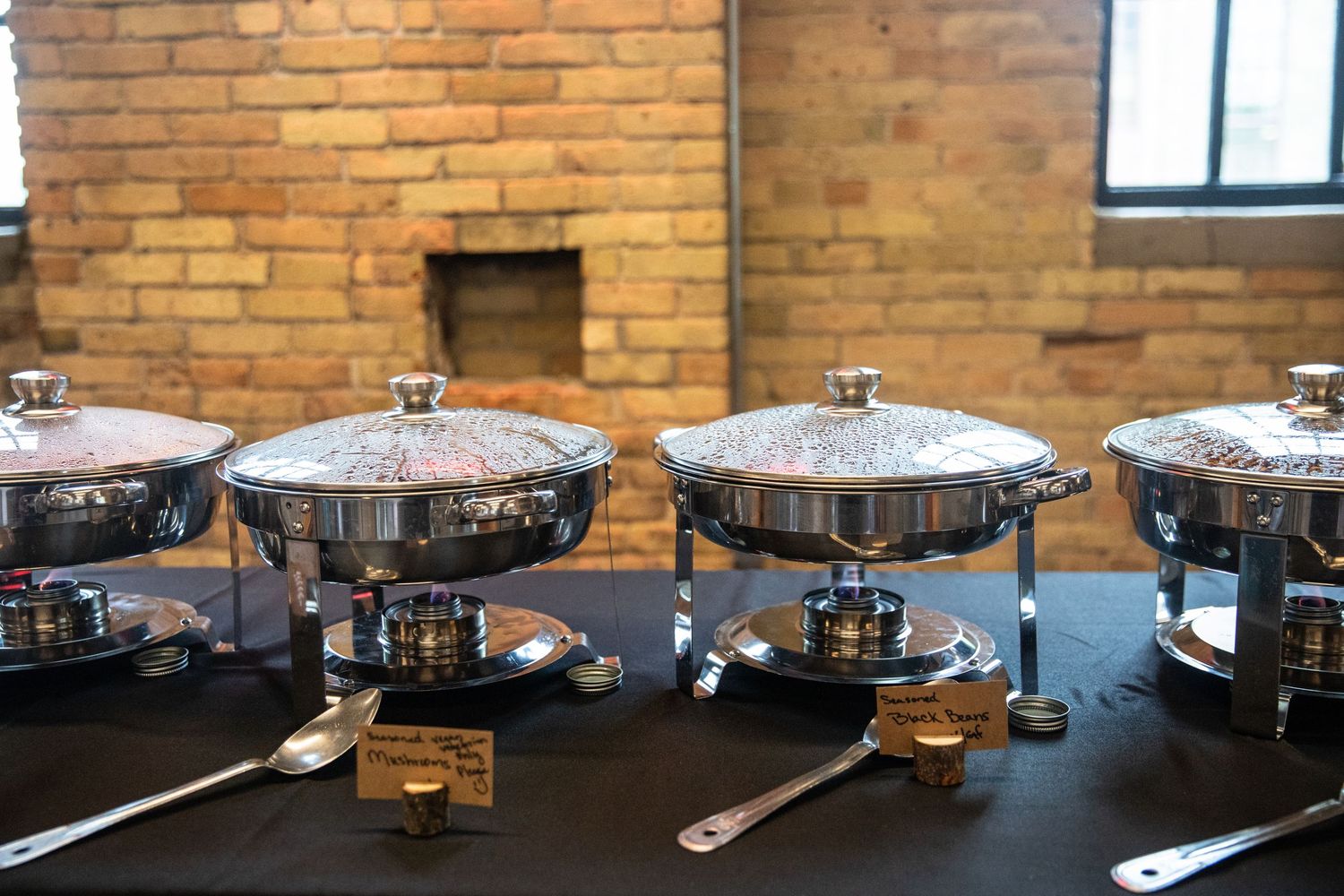 Row of stainless steel chafing dishes lined up on a black tablecloth against exposed brick wall background.