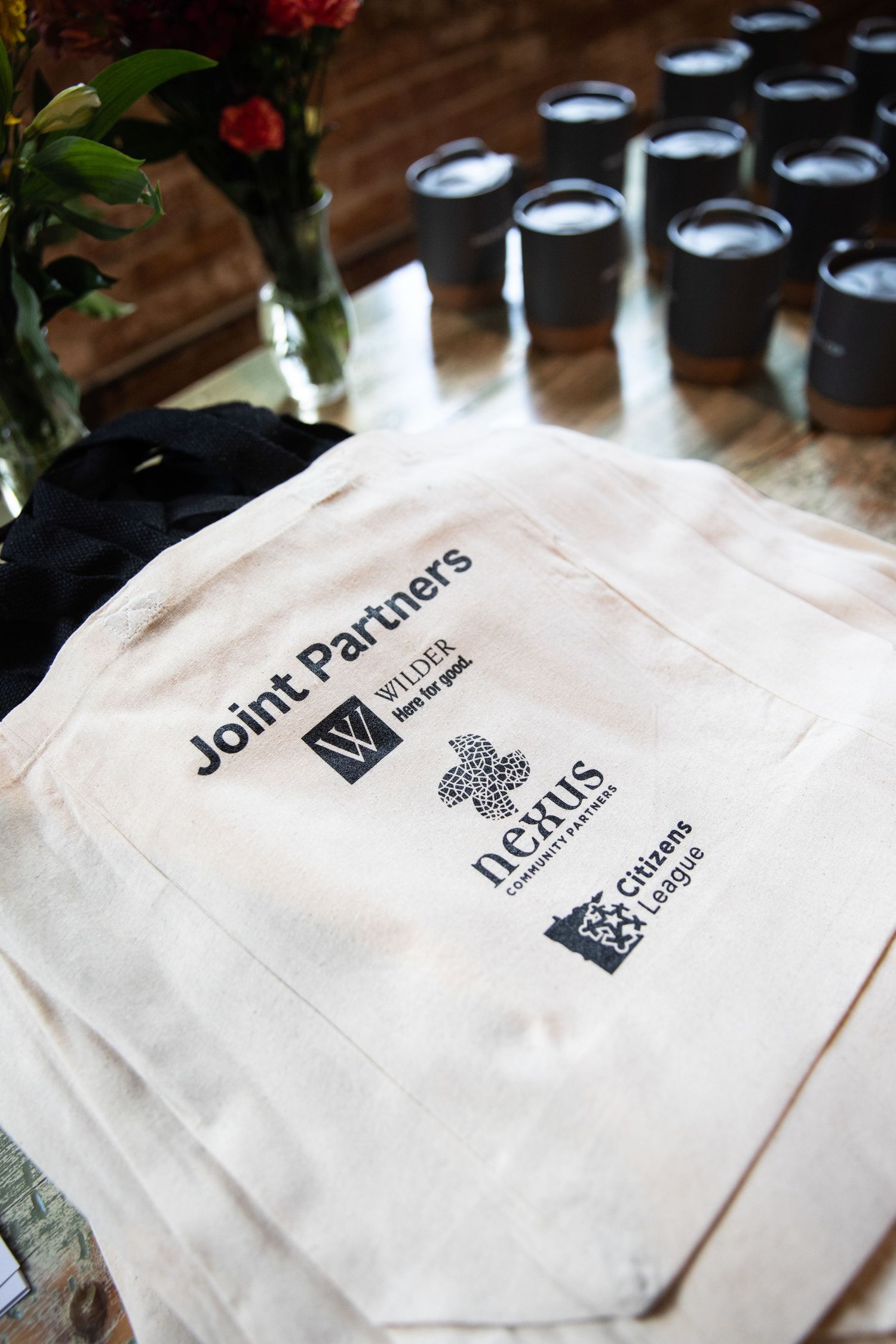 A white canvas bag with Joint Partners branding and multiple company logos sit on a wooden surface beside wine glasses.