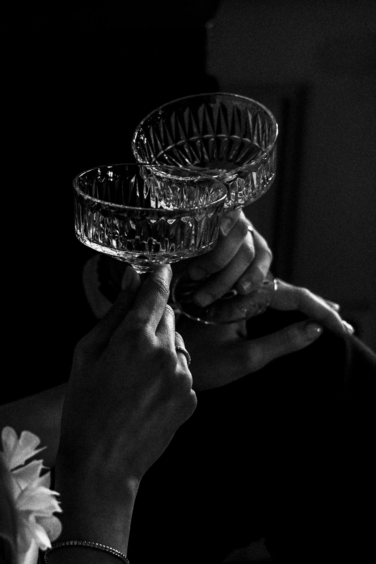 Two hands raise crystal champagne glasses in a toast against a dark background in dramatic black and white lighting.