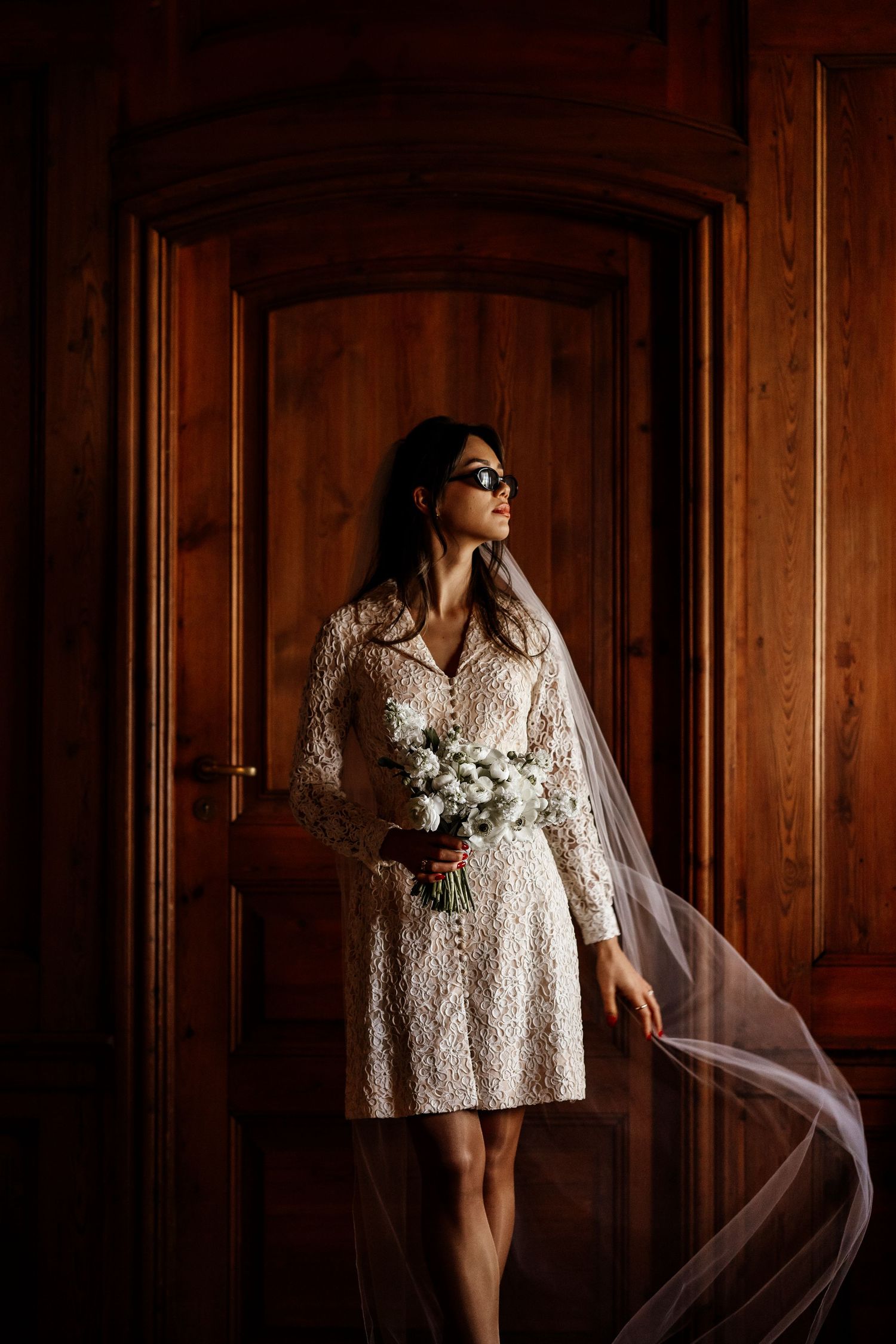 A bride in a short lace wedding dress and veil holds a white bouquet while standing against wooden doors.