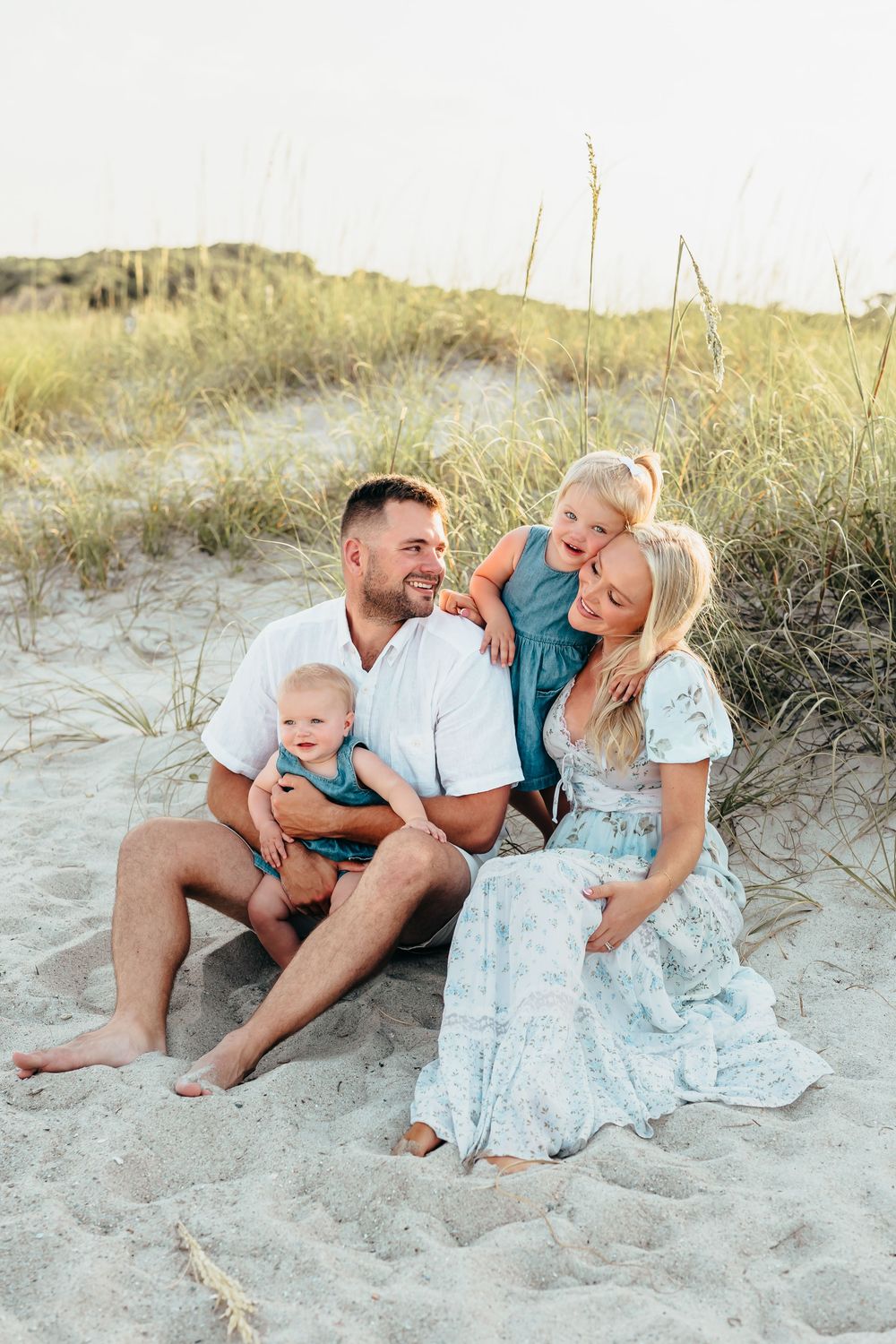 A happy family shares a joyful moment together sitting on sand dunes at the beach during sunset. Wilmington NC.