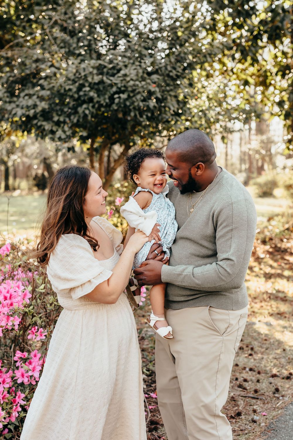 Candid family moments captured in outdoor setting with white and beige outfits near pink flowers.