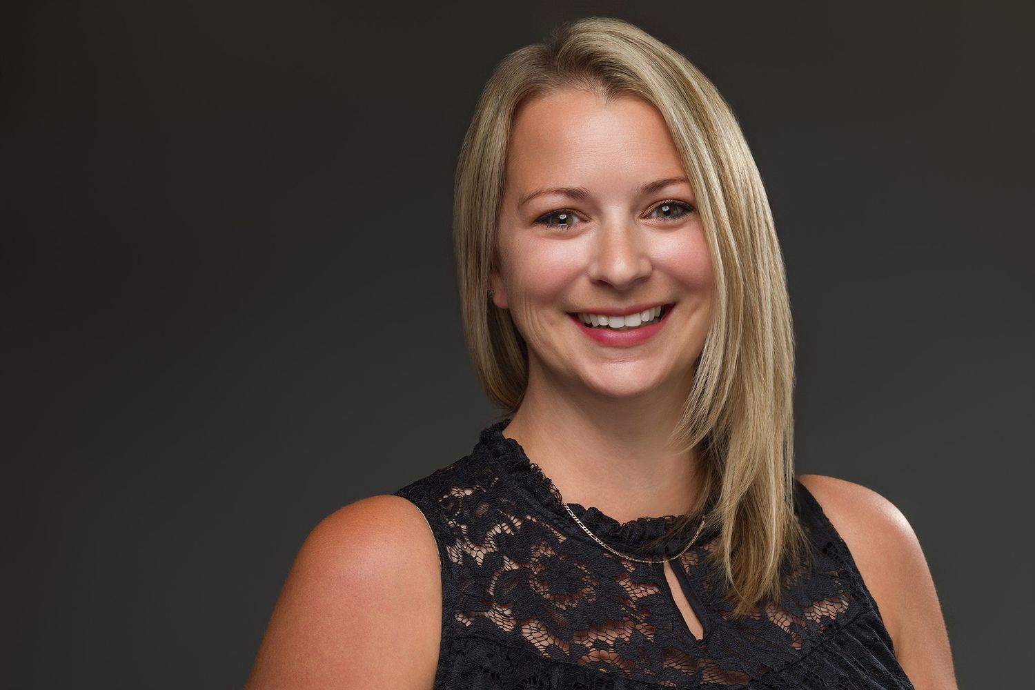 Professional headshot of a blonde person in a black lace top against a dark background.