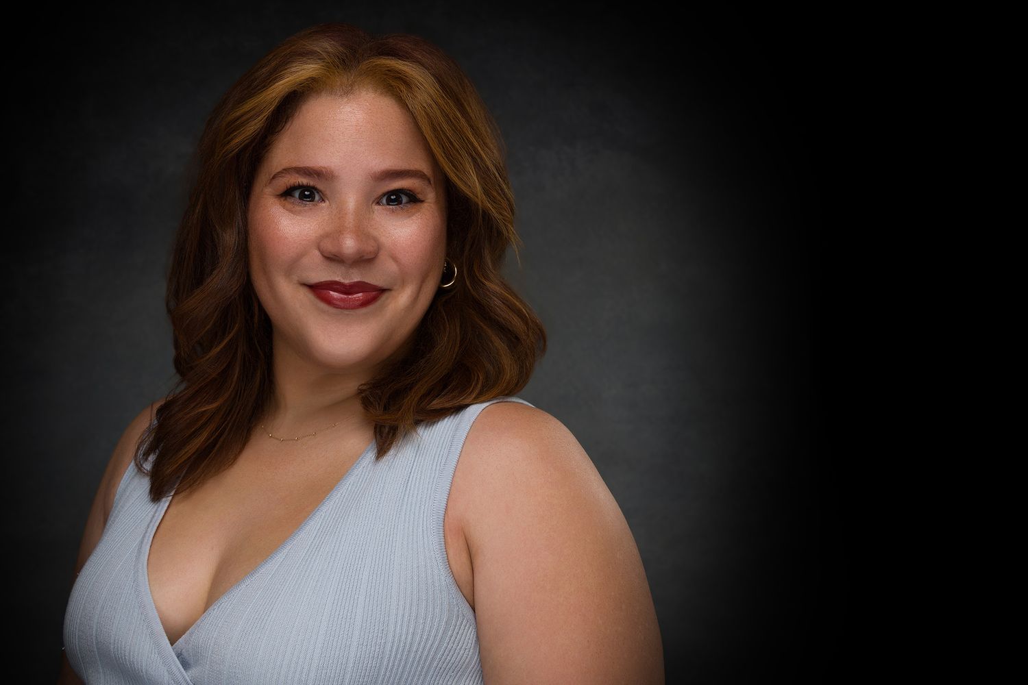 Studio portrait of a smiling woman with brown wavy hair wearing a white sleeveless top against a dark background.