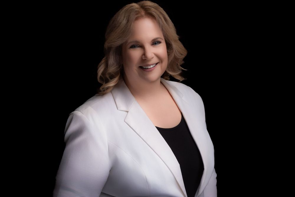 Professional headshot style portrait against black background showing person in white blazer and black top smiling.