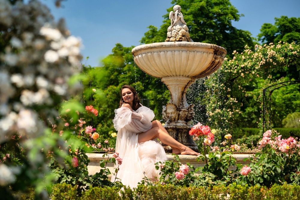A dreamy garden scene with a flowing pink dress against a ornate fountain surrounded by blooming roses and lush greenery.