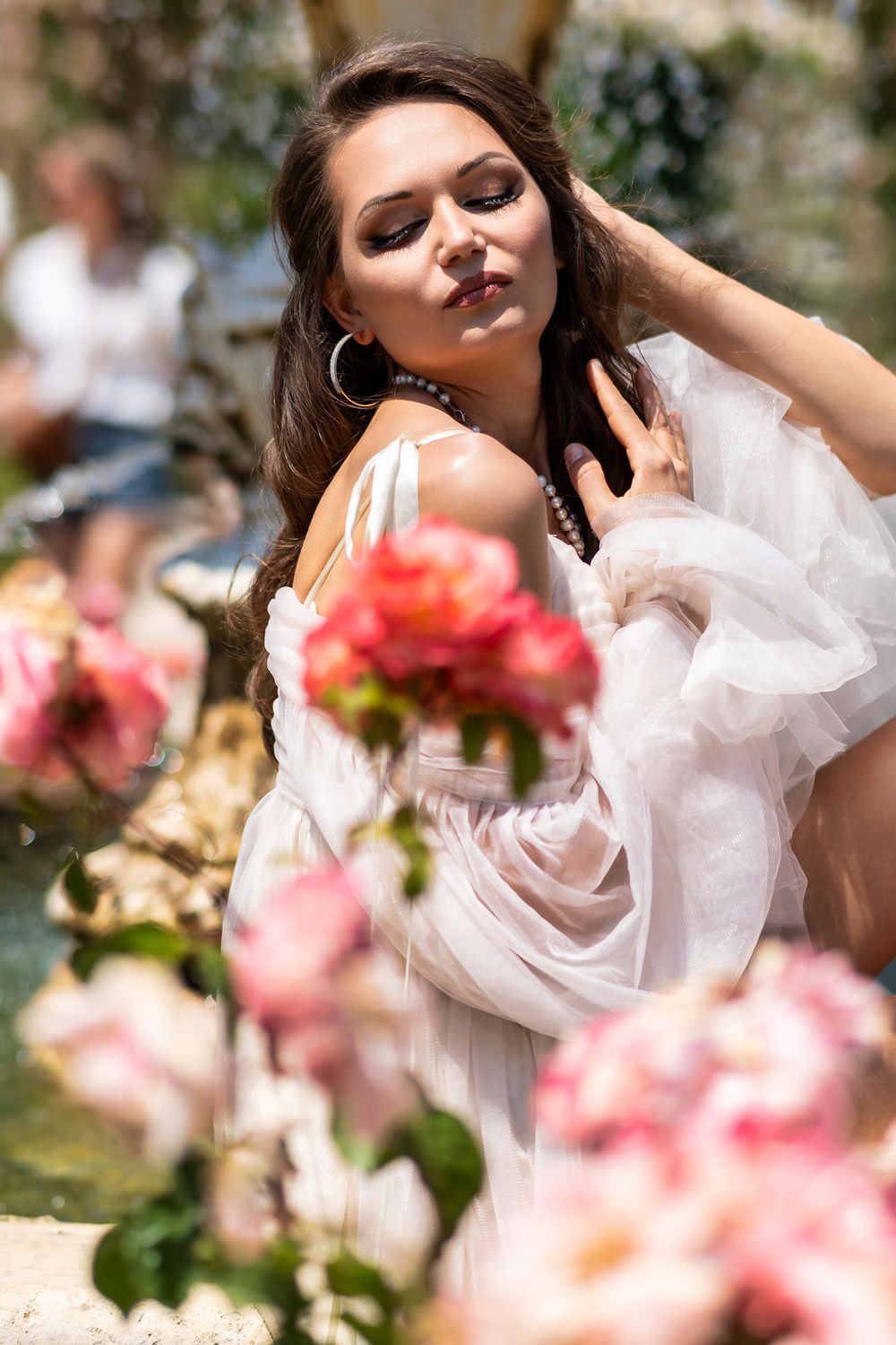 Romantic portrait in a white flowy dress amid blooming pink roses in a sunlit garden setting.