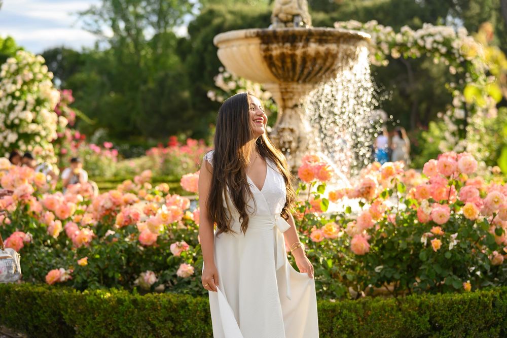 A white-dressed figure stands near an ornate fountain in a blooming rose garden filled with peach-colored flowers.