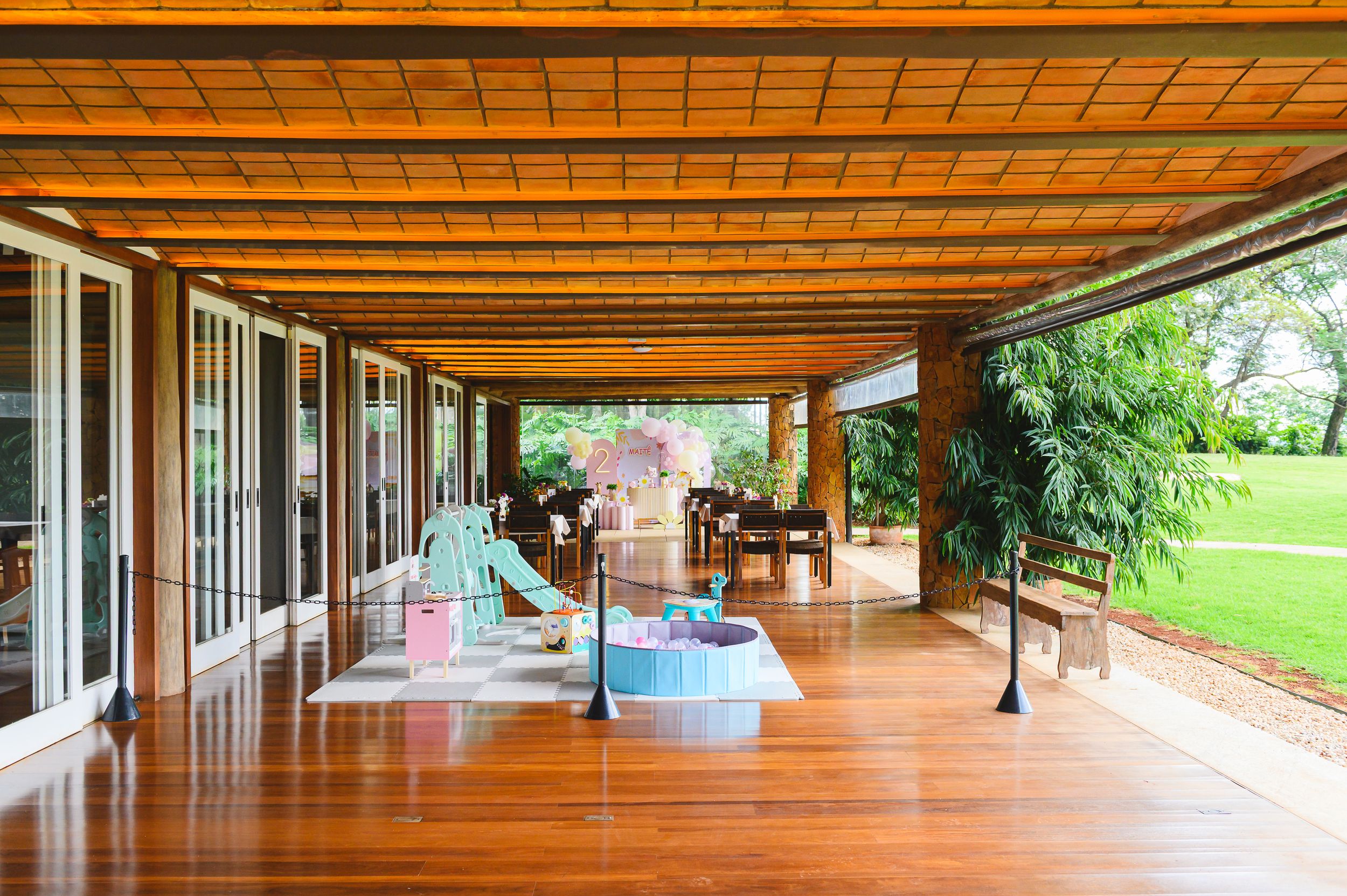 A modern covered porch with wooden decking, turquoise chairs, and tropical plants overlooking a grassy lawn.