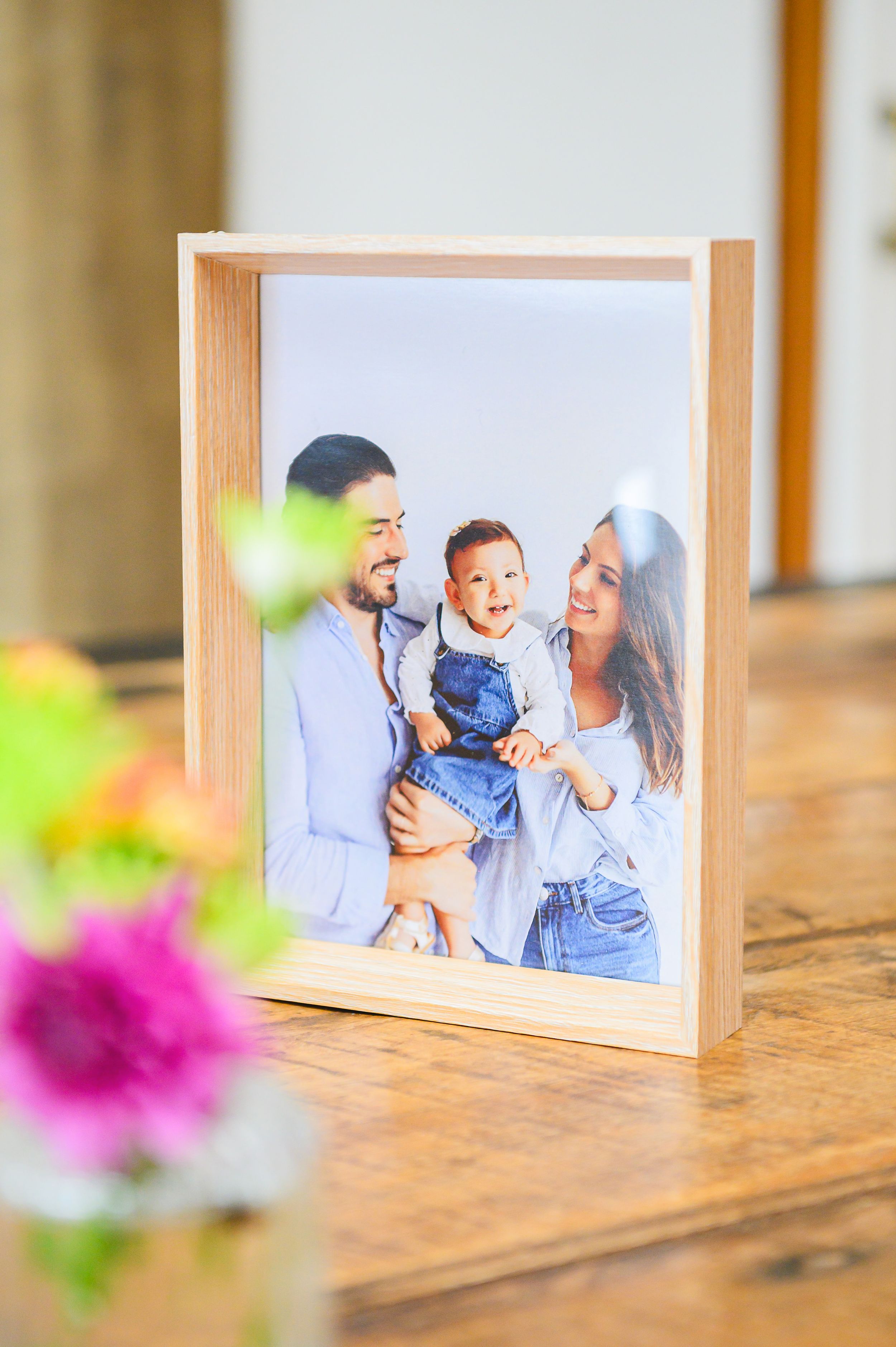 Natural wooden photo frame with a family portrait displayed on a wooden surface with blurred flowers in foreground.