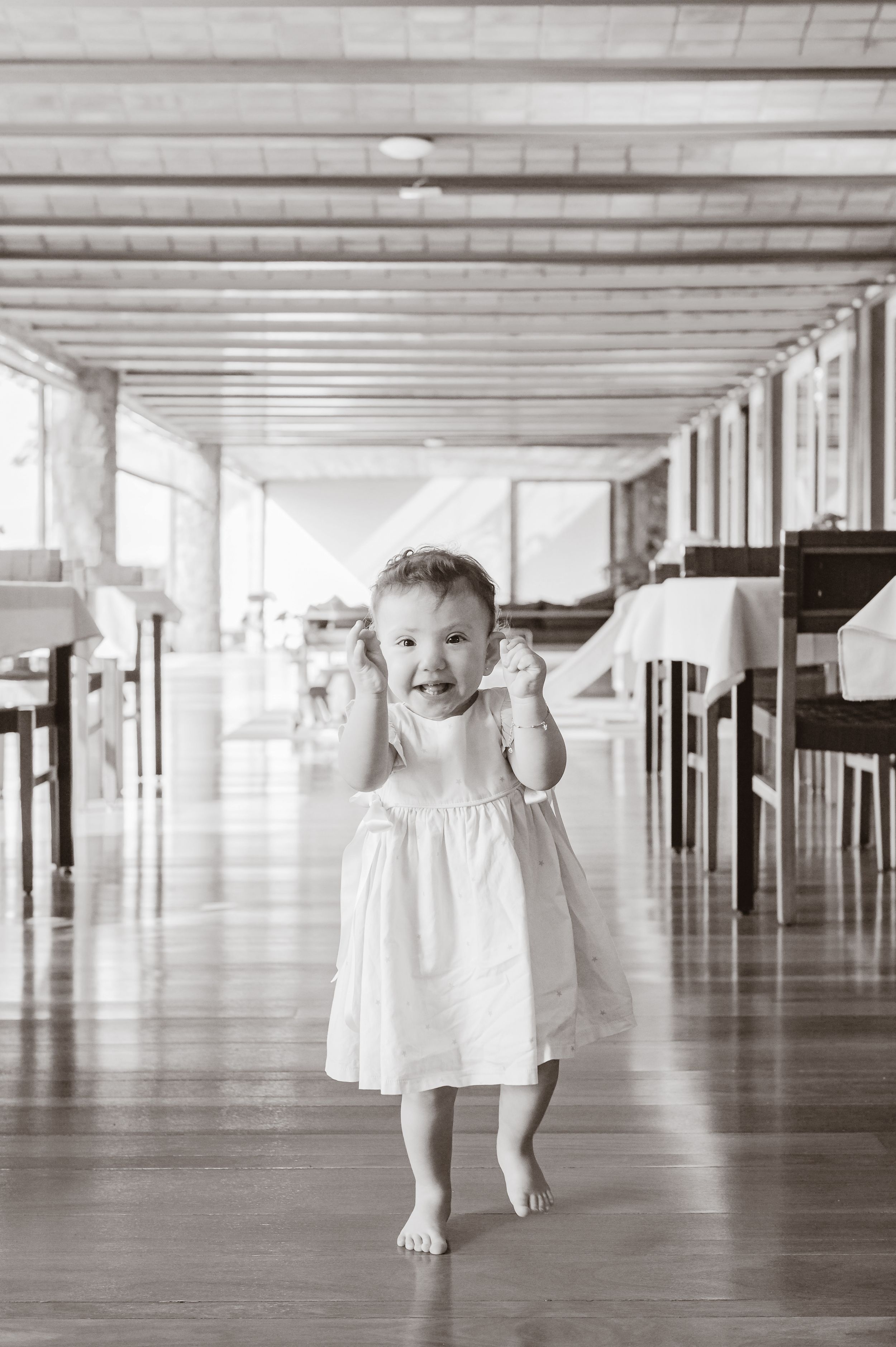 Young child wearing a white dress runs barefoot through a long covered corridor while expressing joy.