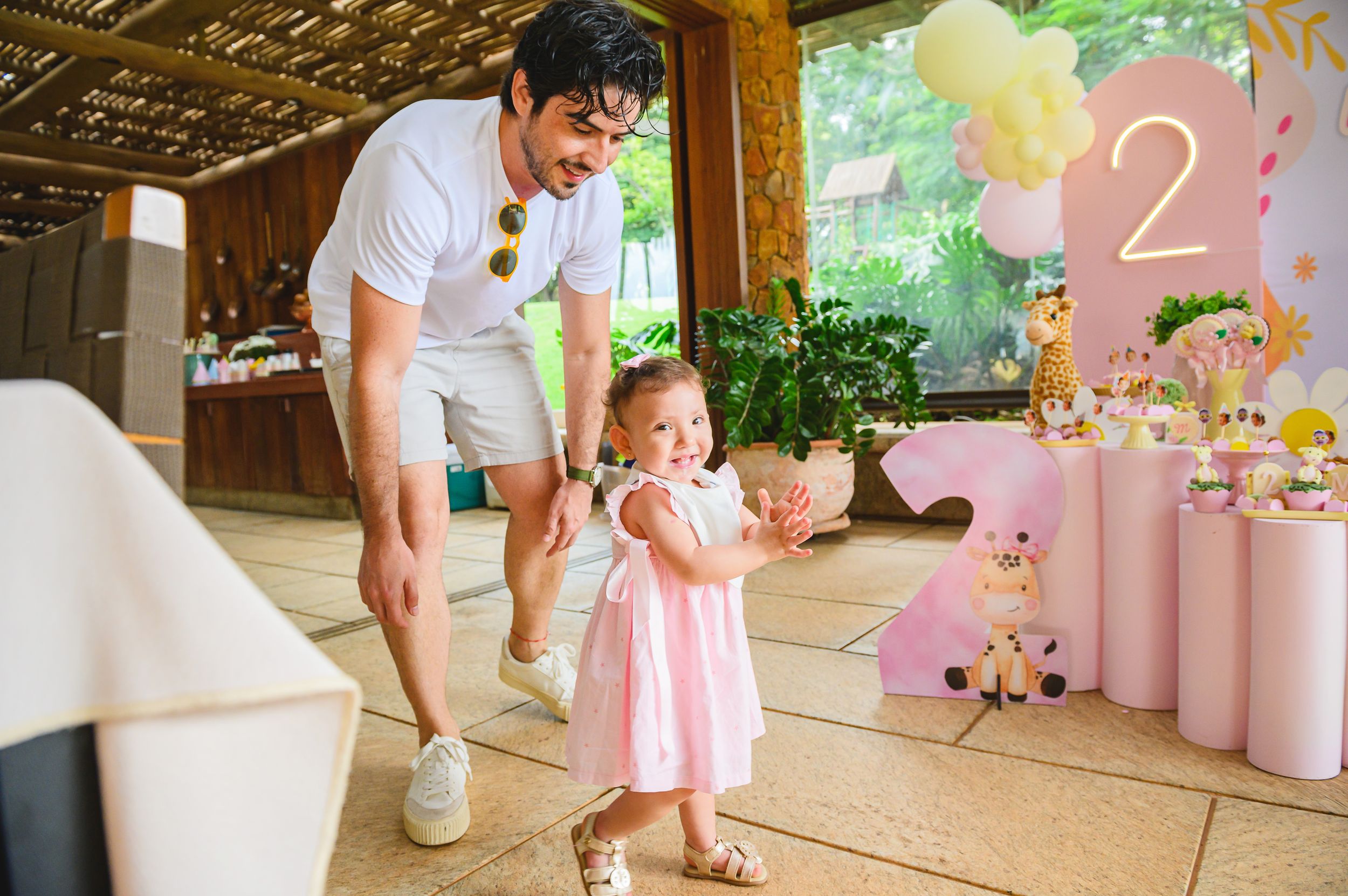 A toddler in a pink dress explores a decorated party venue with soft pink decorations and balloons for a birthday celebration.