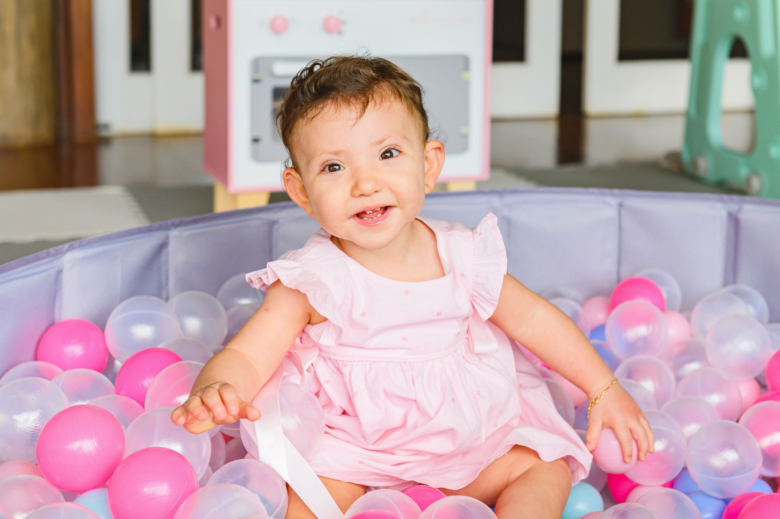 Happy child in pink dress sits in ball pit filled with pink and purple balloons during playtime.