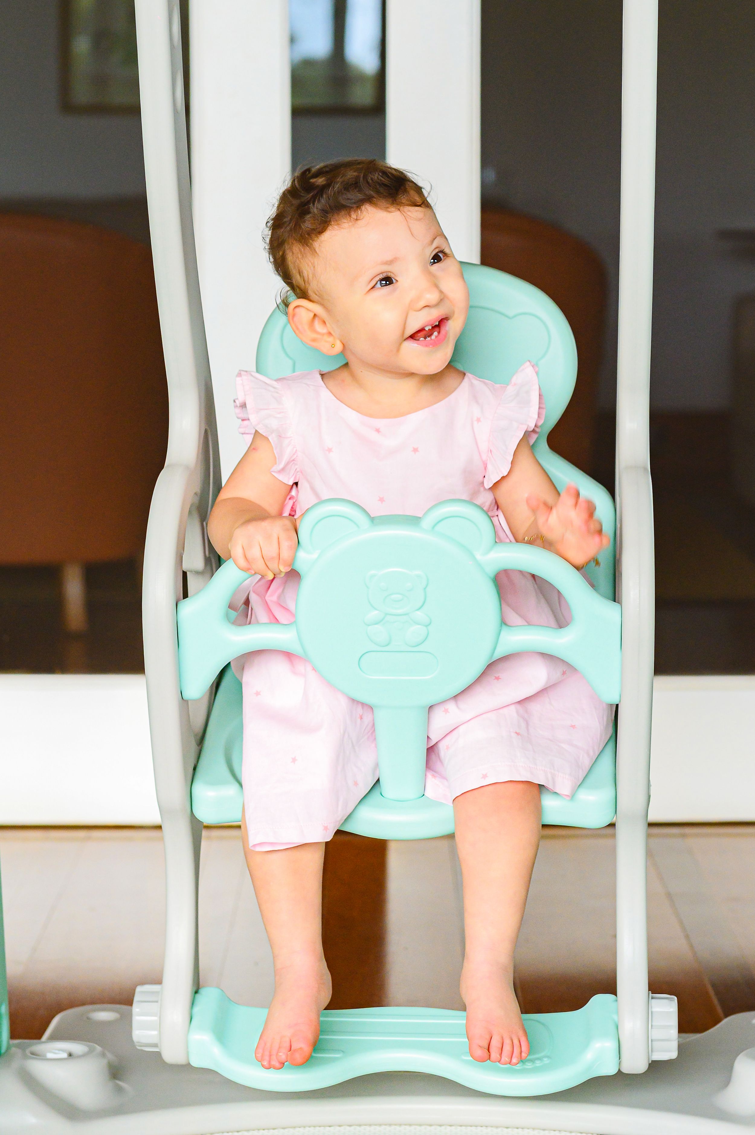 Happy baby sitting in a mint green plastic swing wearing pink and white clothing with bare feet.