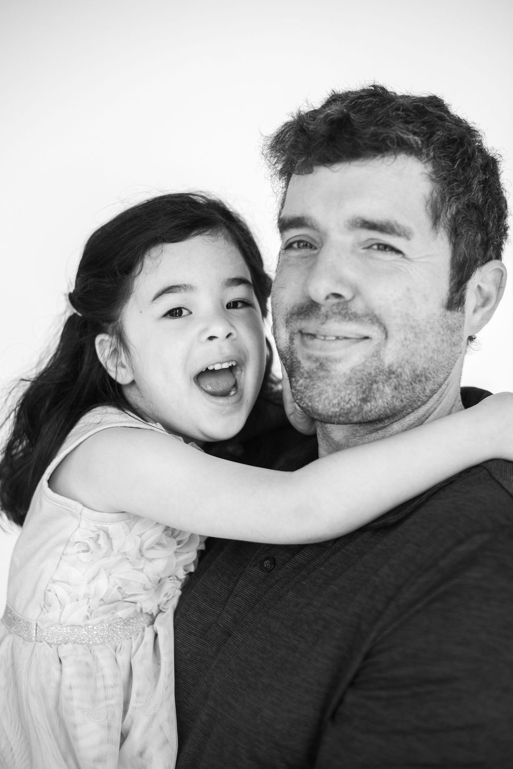 A black and white portrait of a young girl in a light dress hugging and laughing with an adult in a dark shirt.