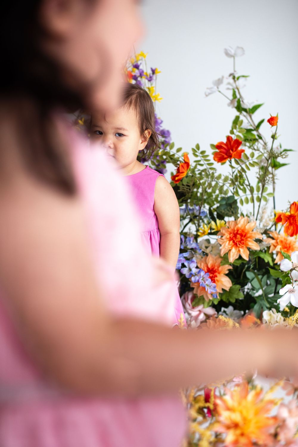 A child in a pink dress peers at colorful summer flowers and greenery in an artistic indoor portrait.
