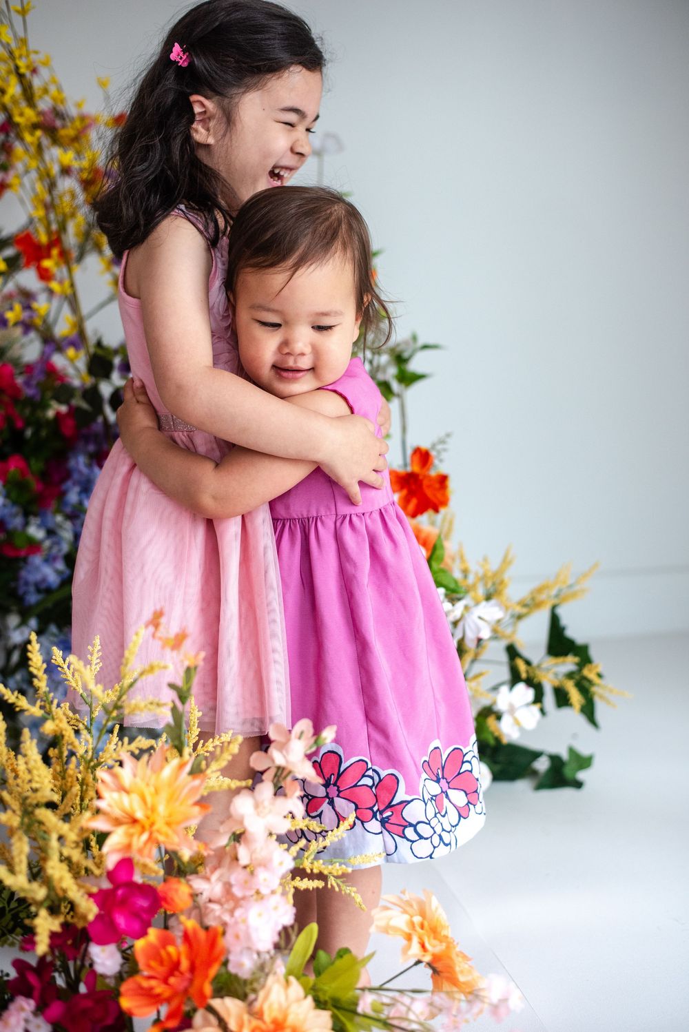 Two adorable children in pink dresses share a warm embrace surrounded by colorful spring flowers and foliage. Taken at Studio 428 in va