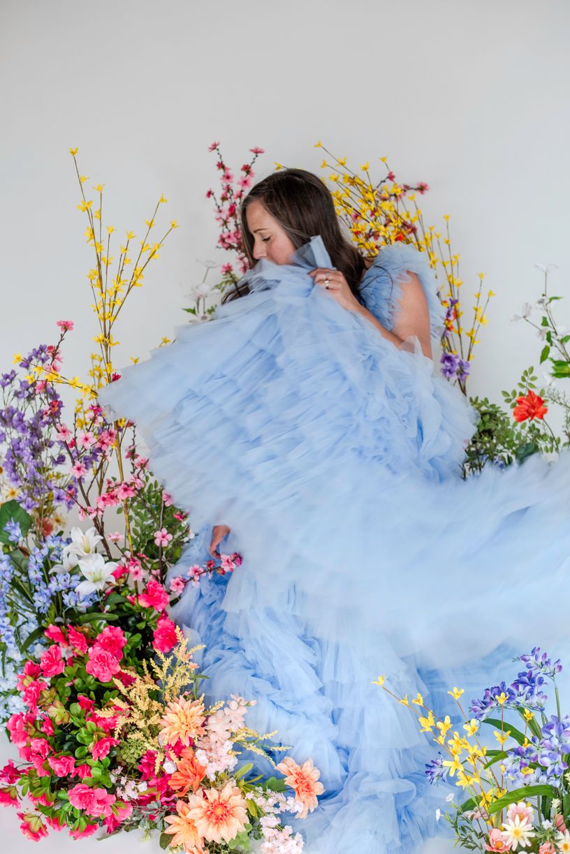 Dreamy portrait with flowing light blue tulle gown surrounded by vibrant spring flowers and yellow forsythia branches.