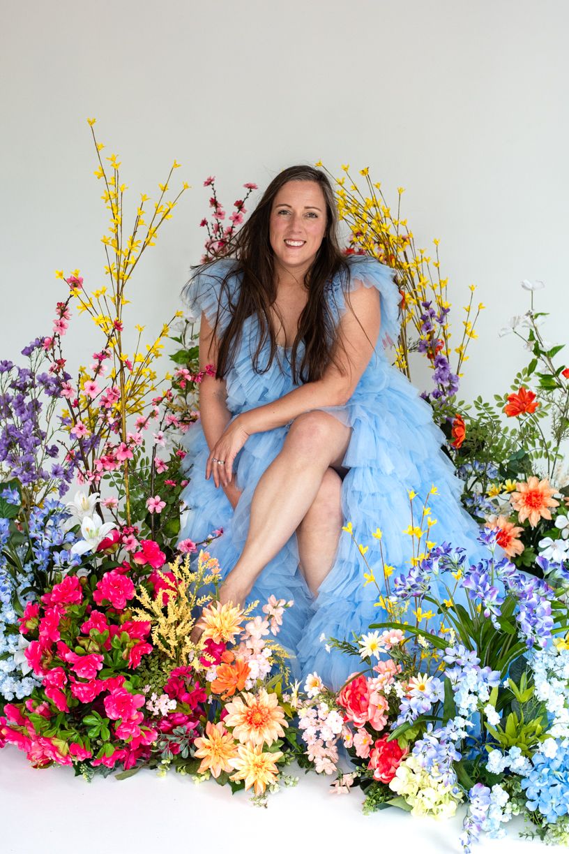 A person in a light blue dress poses among a vibrant display of colorful spring flowers including forsythia and delphiniums.