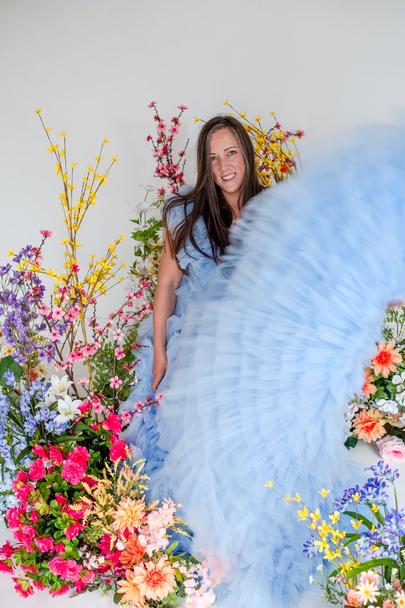 A flowing light blue tulle gown surrounded by vibrant spring flowers and colorful floral arrangements against a white background.