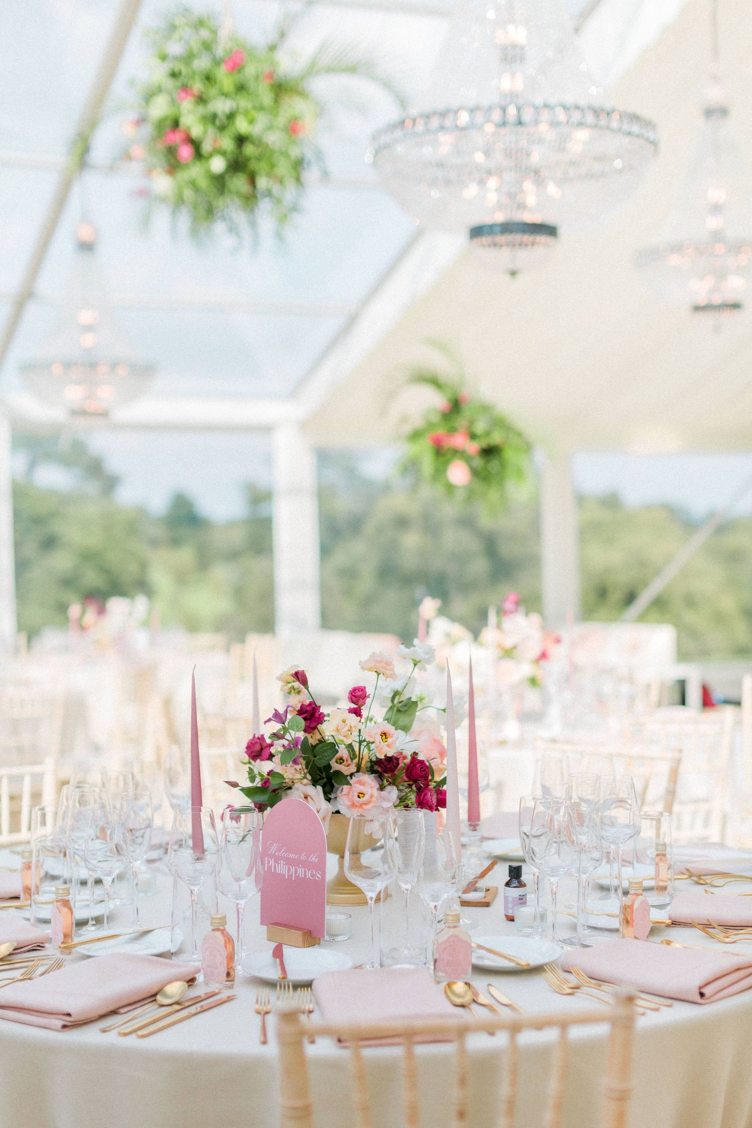 Elegant wedding reception table with pink floral centerpiece, crystal chandelier and hanging plants in glass marquee at Sezincote Cotswolds
