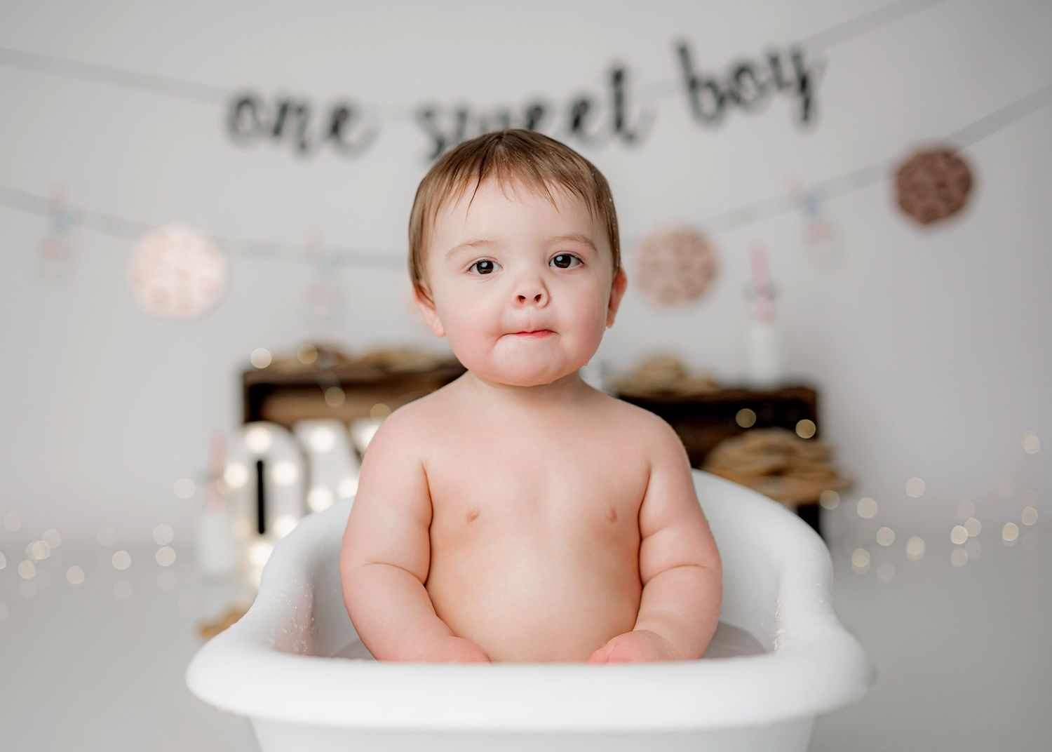 A laughing baby sits in a white bathtub under a banner and string lights with delicate decorative snowflakes.