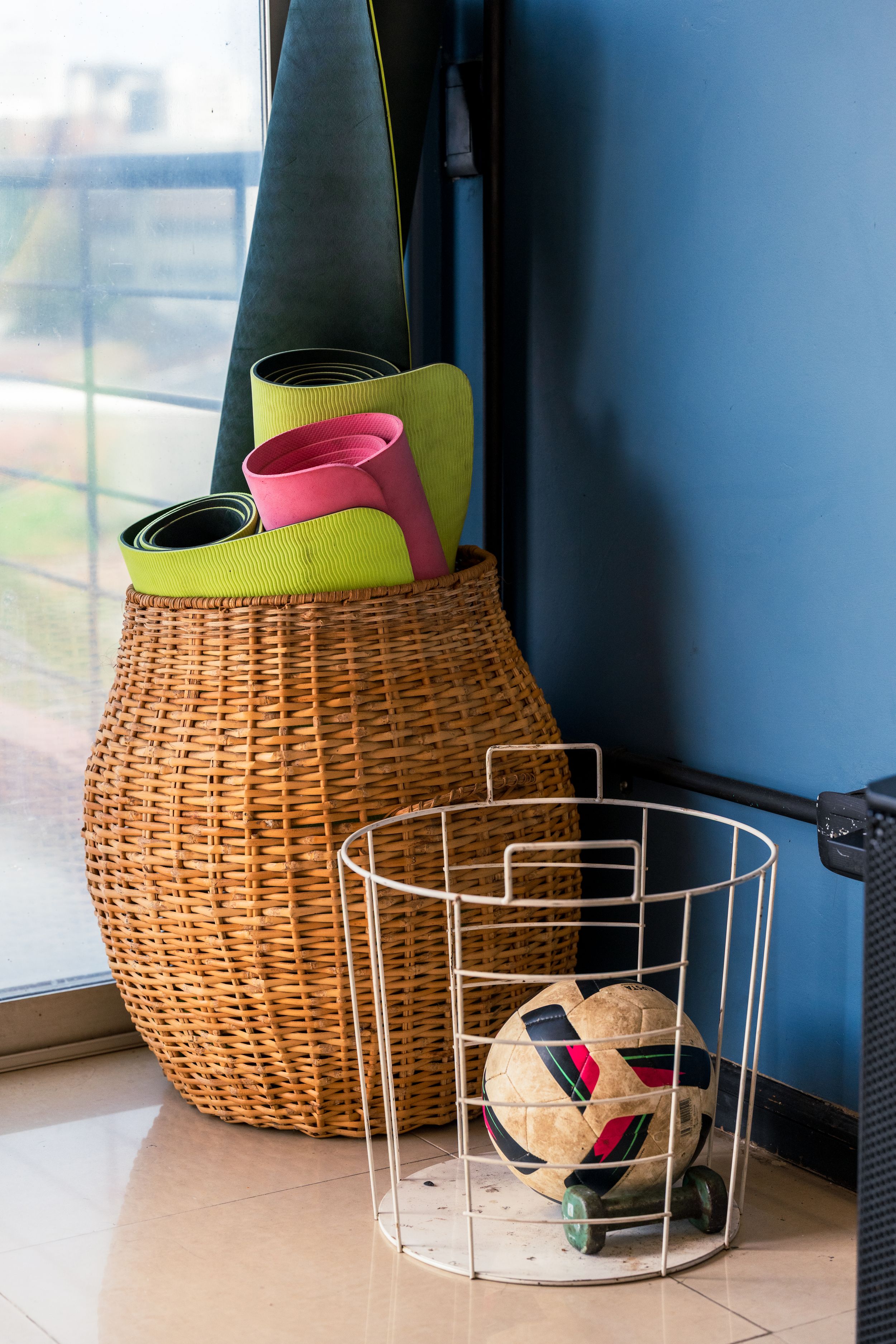 Wicker basket with yoga mats and a metal wire basket containing a soccer ball near a window.