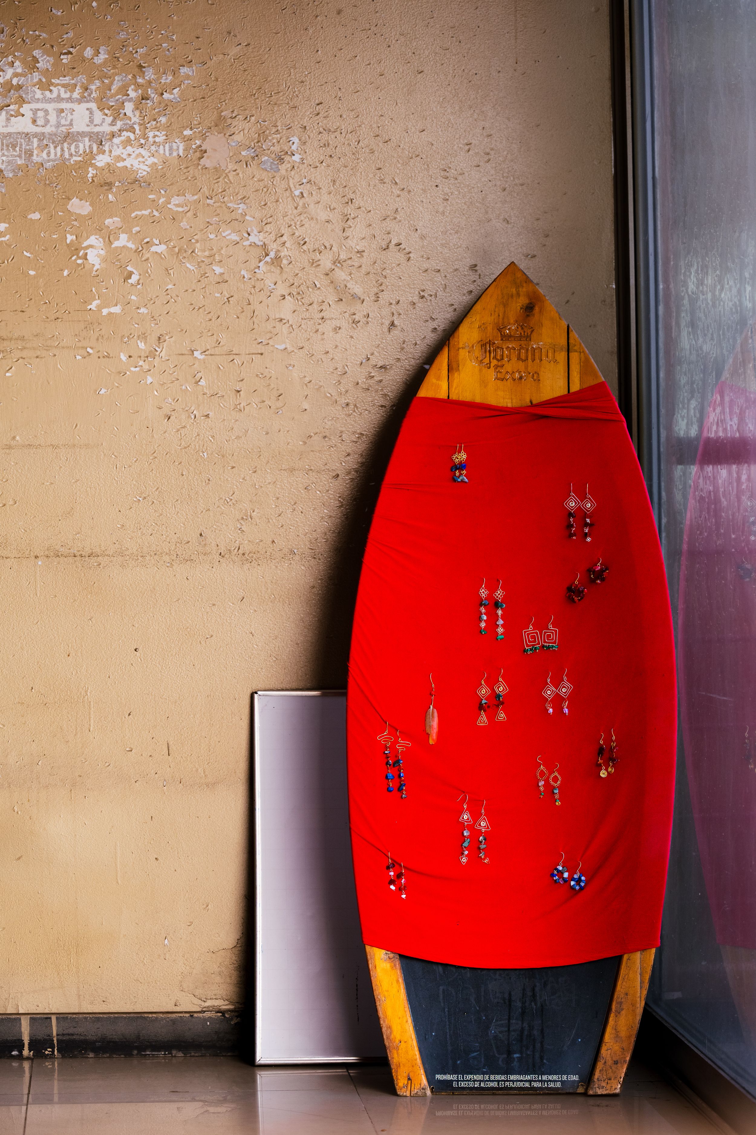 A vintage red surfboard with decorative pins leaning against a beige wall in a corner.
