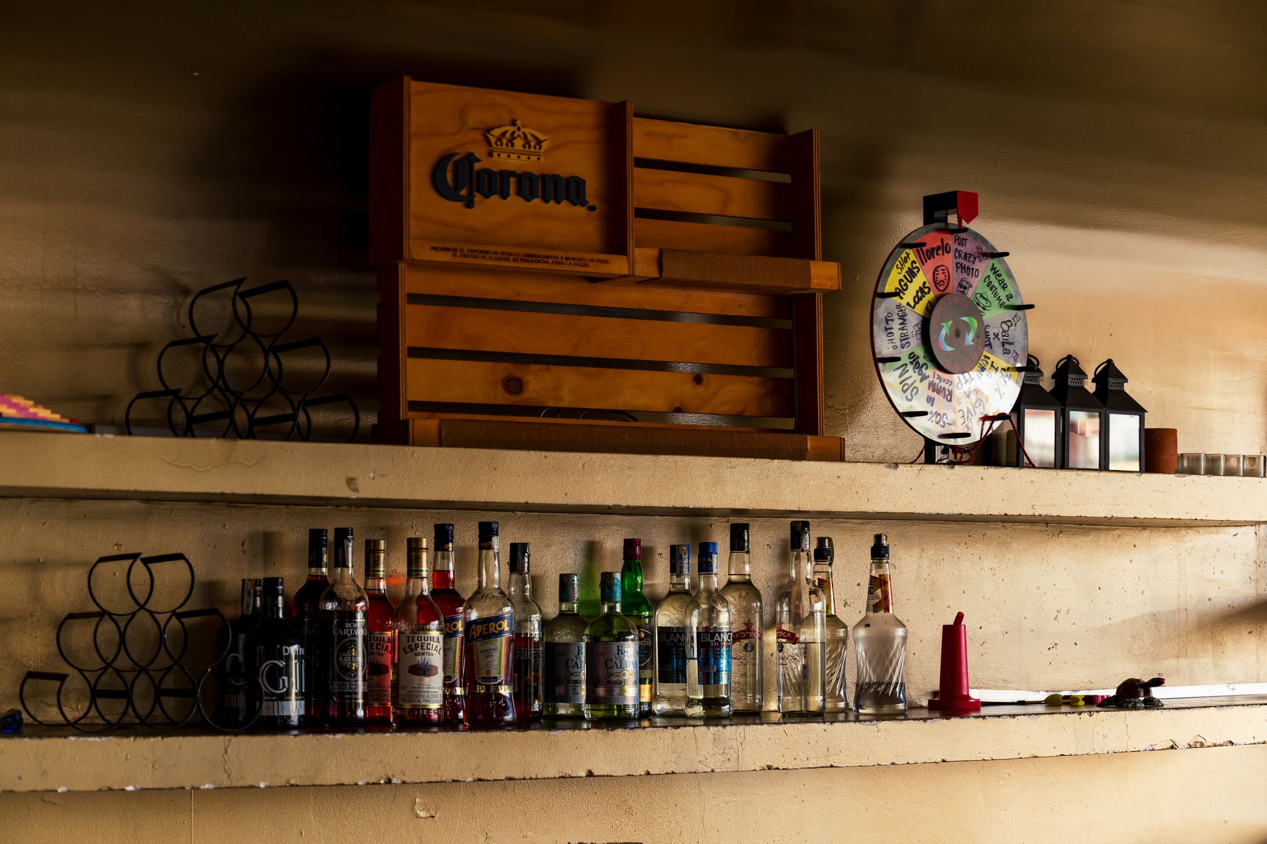 Two wooden shelves display a collection of liquor bottles and wine glasses in a dimly lit bar setting.