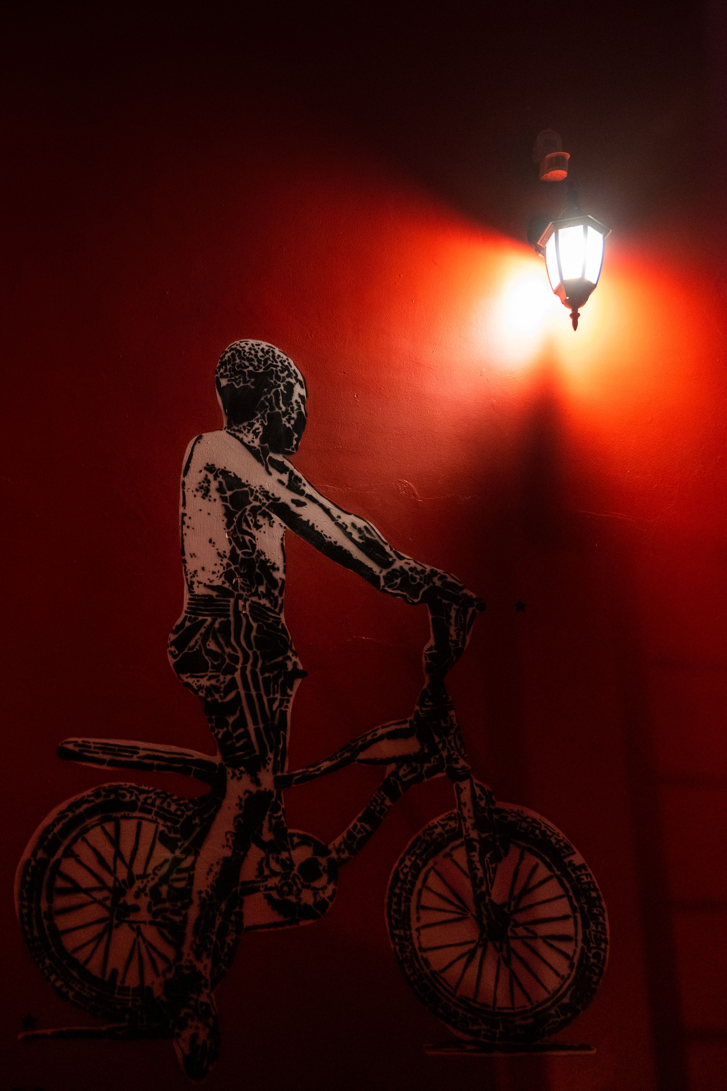 A silhouette of a person with a bicycle stands under a glowing red streetlamp creating a dramatic atmospheric scene.