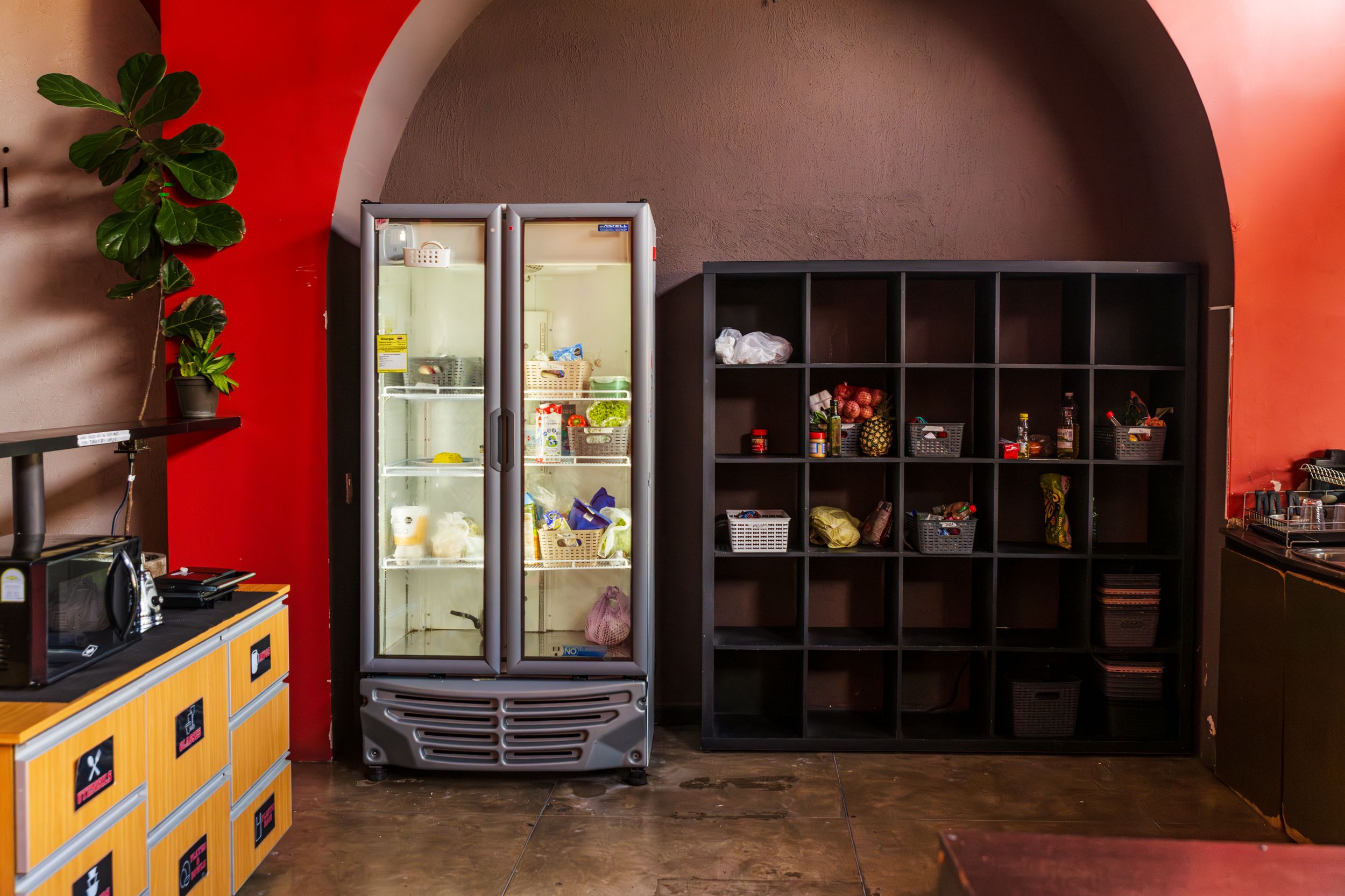 Interior of a cafe with red walls, dark wooden storage cubes, and a glass refrigerator display case in an arched alcove.